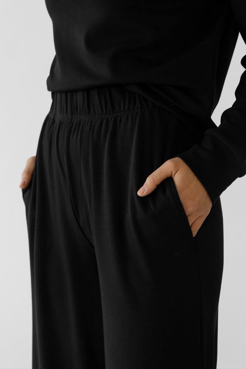 A model is wearing the Women's Ultra-Soft Bamboo Wide Leg Pull On Pant & Pullover Set by Cozy Earth. The outfit, featuring a black hoodie and pants with hands in pockets, exudes a casual and comfortable vibe against a plain white background.
