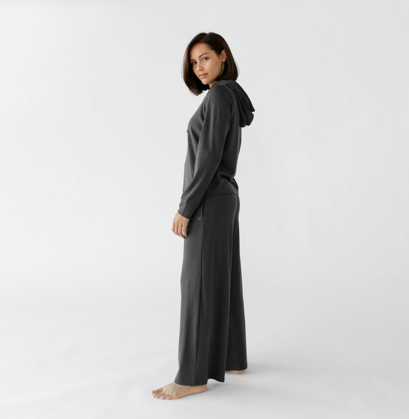 A person with shoulder-length hair stands barefoot in a minimalist, white setting, wearing Cozy Earth's Women's Ultra-Soft Bamboo Wide Leg Pull On Pant & Hoodie Set in dark hues, looking over their shoulder. 