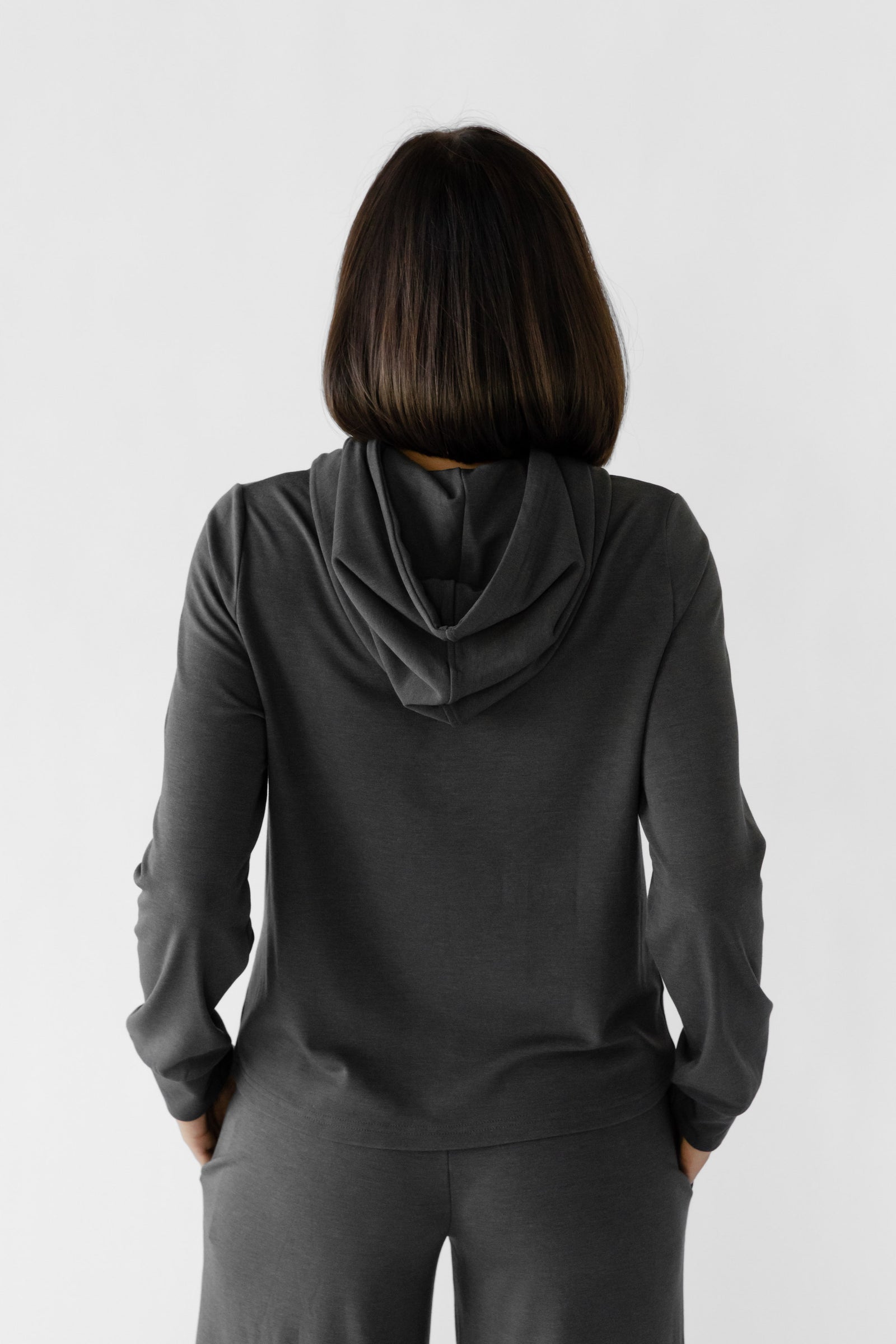 A person with shoulder-length brown hair stands facing away, wearing Cozy Earth's Women's Ultra-Soft Bamboo Wide Leg Pull On Pant & Hoodie Set in dark gray against a plain white background. 