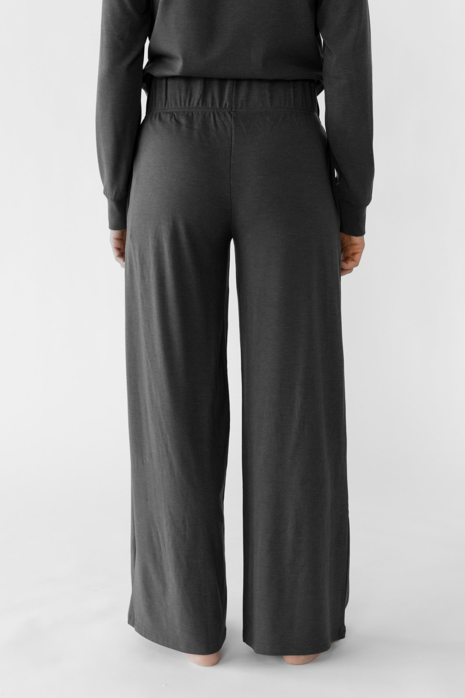 A person is standing facing away, wearing the Women's Ultra-Soft Bamboo Wide Leg Pull On Pant & Hoodie Set by Cozy Earth in dark gray against a plain white background. 