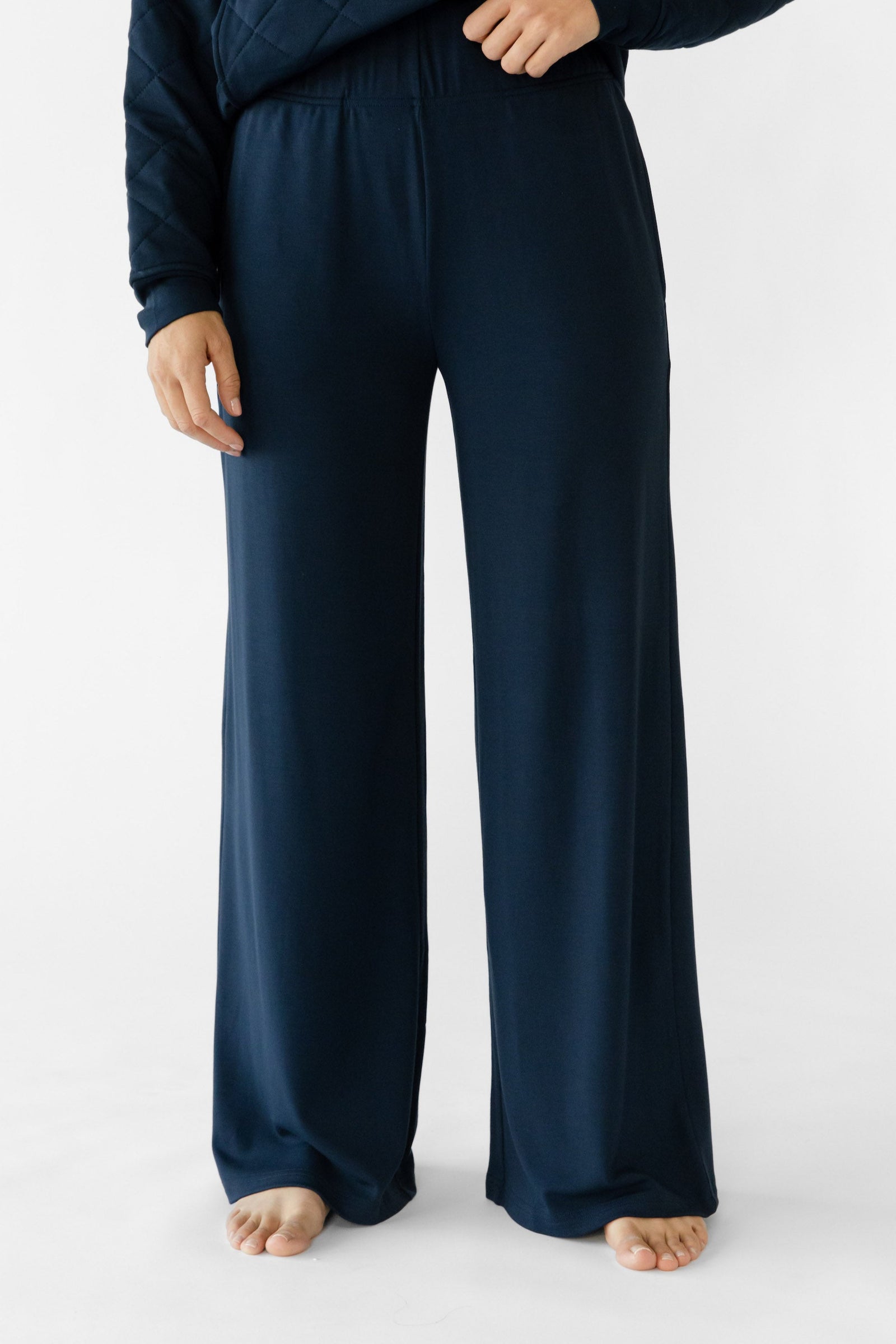 A person in a dark blue Cozy Earth Women's Ultra-Soft Bamboo Wide Leg Pull On Pant & Pullover Set stands barefoot on a white background, with their left hand resting on their hip, exemplifying comfort and casual style. 