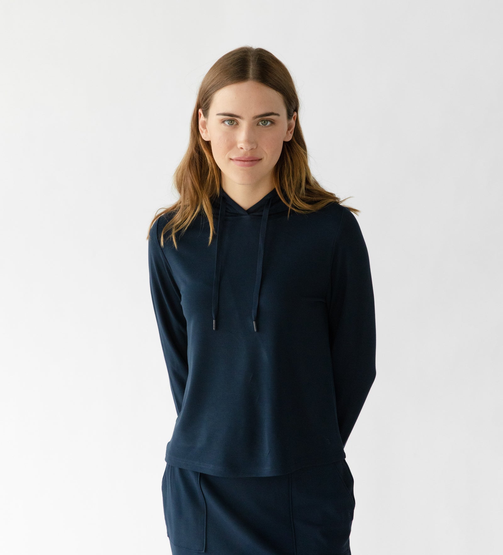 A person with long brown hair is wearing the dark blue Cozy Earth Women's Ultra-Soft Bamboo Wide Leg Pull On Pant & Hoodie Set against a plain white background, hands behind their back, with a neutral expression. 