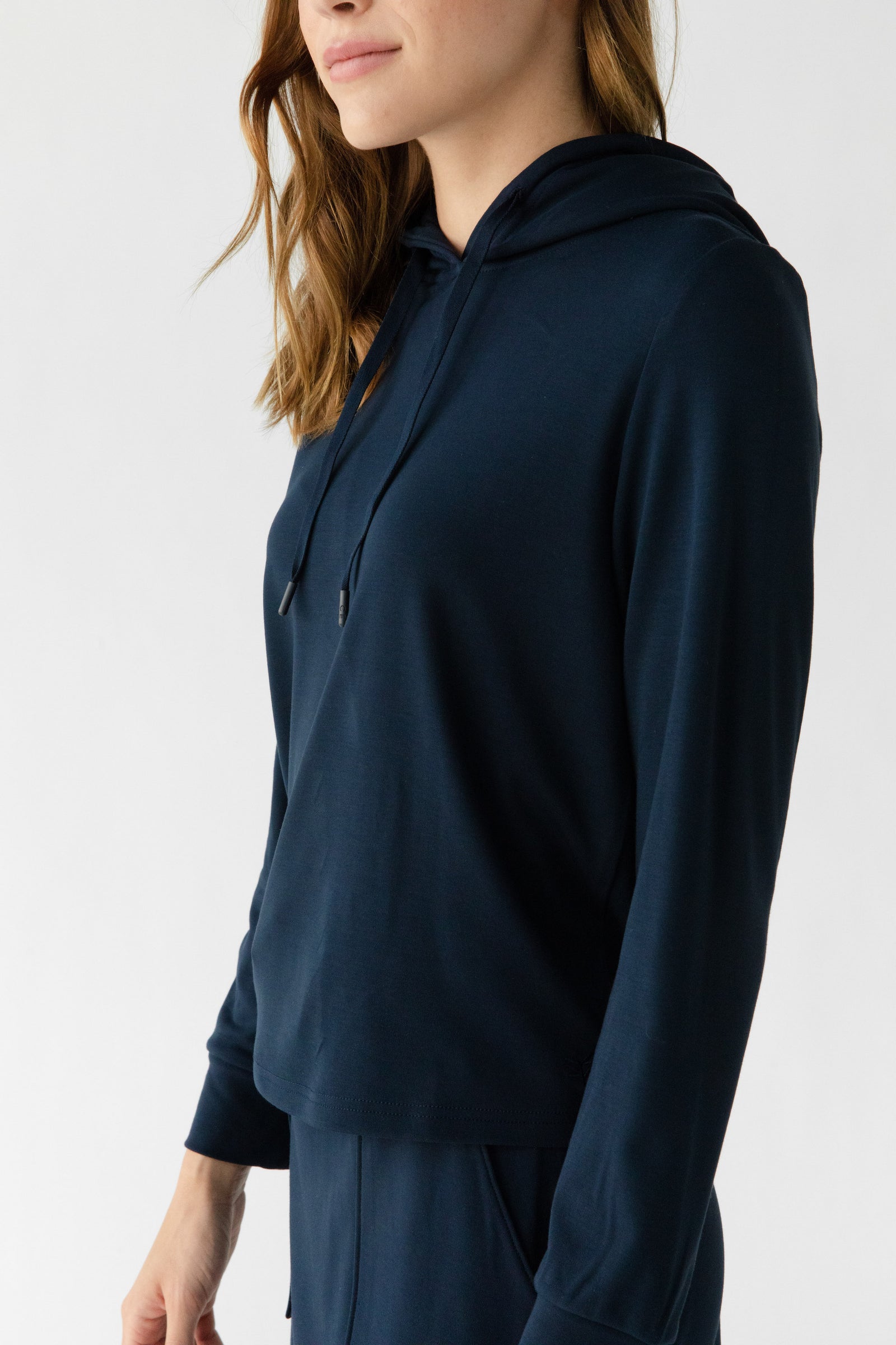 A woman models the Women's Ultra-Soft Bamboo Wide Leg Pull On Pant & Hoodie Set by Cozy Earth in dark blue. She stands against a plain white background, her long, wavy hair partially obscuring her face as she gazes to the side. 