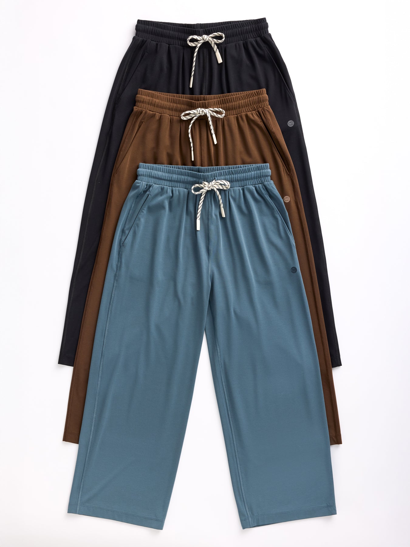 Four pairs of Cozy Earth's Women's Studio Cropped Wide Leg Pants are stacked against a white background in colors black, brown, dark brown, and blue. Each pair features a drawstring waistband and visible side pockets. 