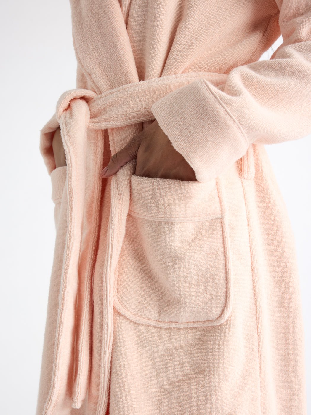 A person wearing the Luxe Bath Robe by Cozy Earth, in soft pink with a tied belt, is shown from shoulders down. One hand is in the front pocket, highlighting its texture and design against a plain white background. 