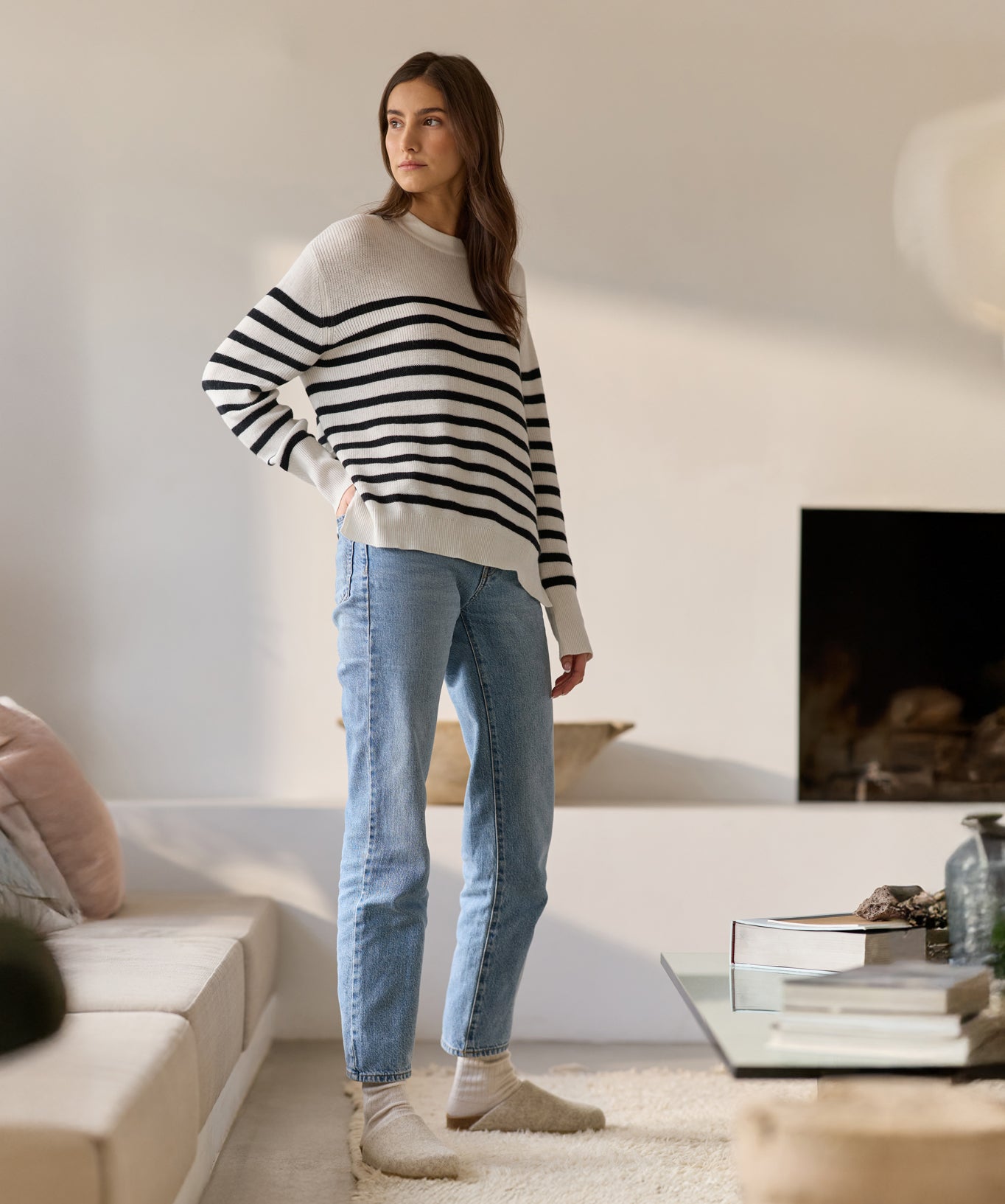 In a modern living room with neutral tones and a cozy atmosphere, a person stands wearing a Cozy Earth's Women's Rowan Sweater, blue jeans, and beige slippers beside a low couch and coffee table, with a lit fireplace in the background. 