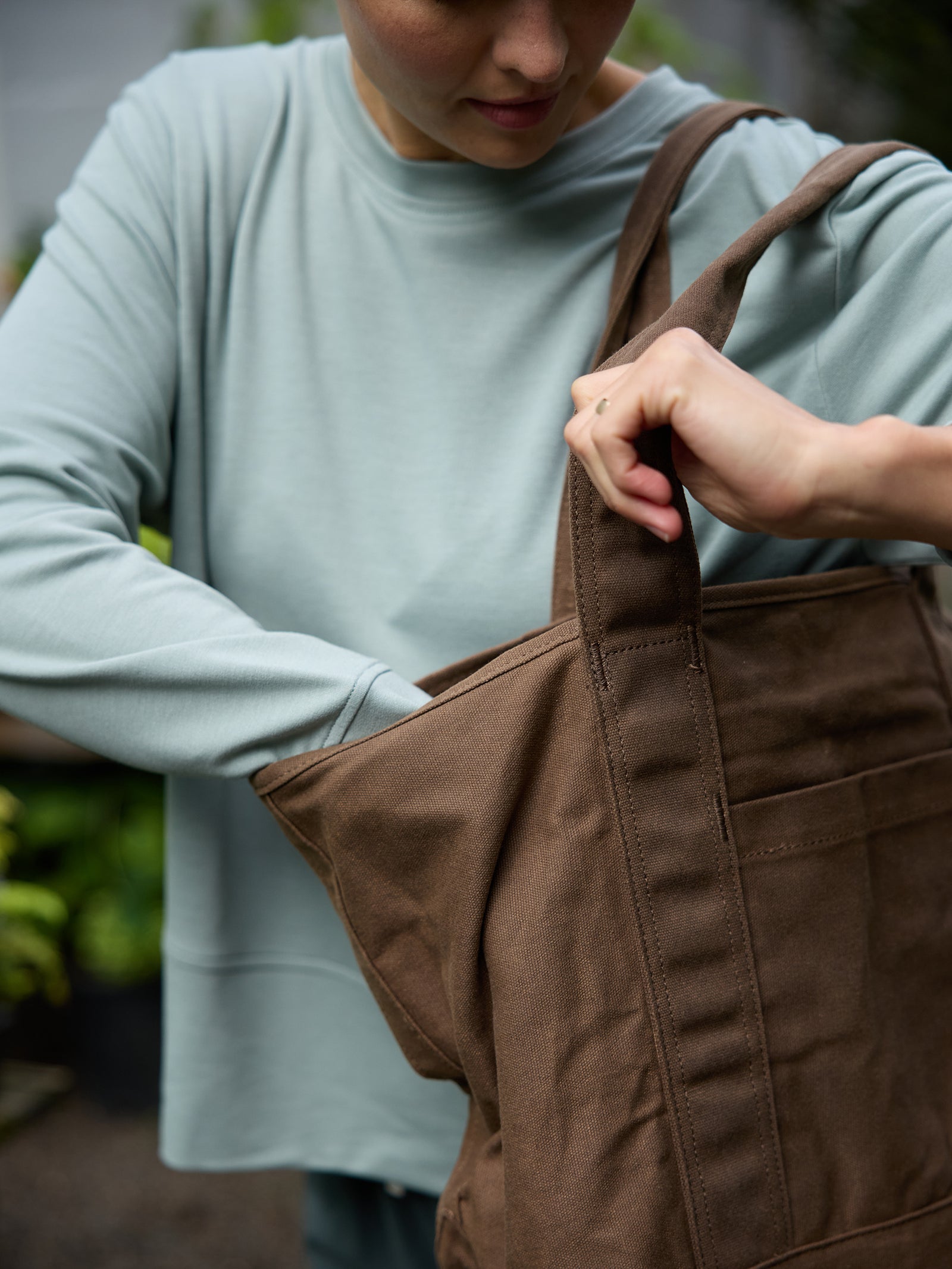 A person wearing a light blue long-sleeve shirt is reaching into a Cozy Earth Waxed Canvas Tote with their right hand. The background is slightly blurred, with some greenery visible. 