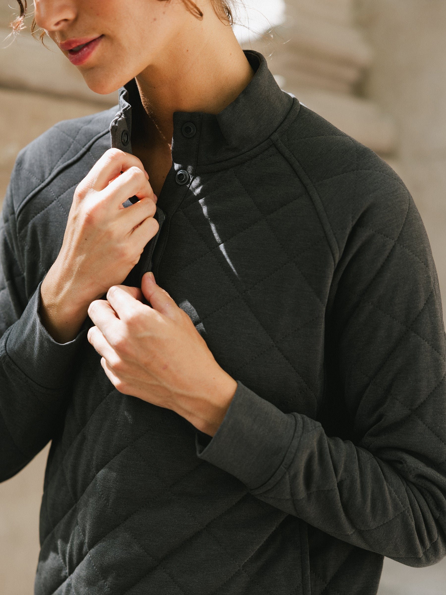 A person with light skin and short wavy hair is adjusting the buttons on their dark-colored Women's Ultra-Soft Bamboo Quilted Snap Pullover from Cozy Earth. They are standing in a well-lit area with a neutral background. 