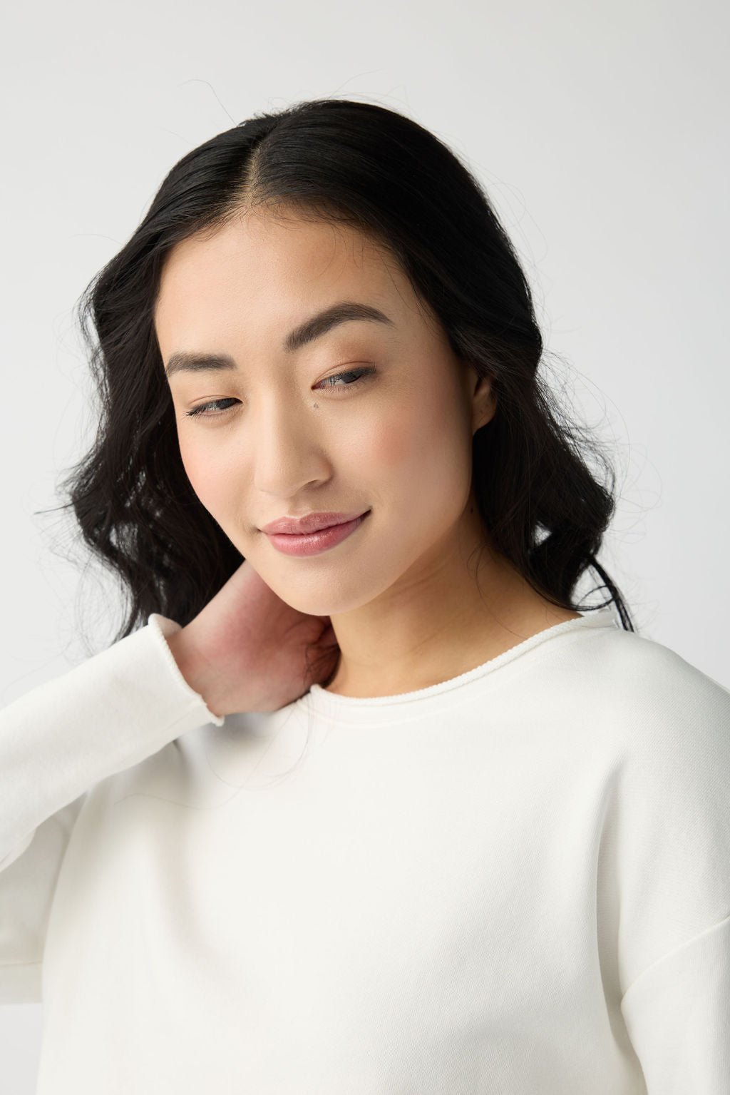 A woman with long, wavy black hair gently touches her neck and smiles softly as she looks down. She is dressed in a white top from the Women's Chelsea Set by Cozy Earth, set against a light gray background. 