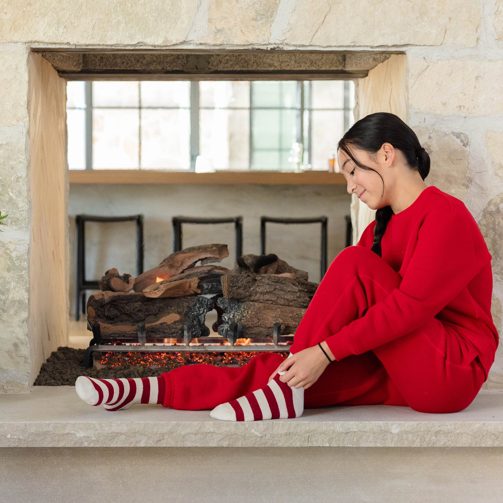 A child wearing Cozy Earth's Kid's CityScape Crewneck and striped socks sits by a stone fireplace, adjusting their sock. The setting is cozy, with logs in the fireplace and a reflective window above. 