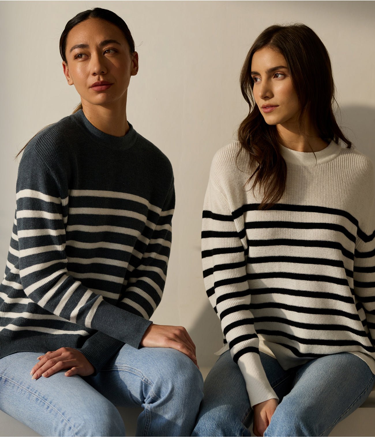 Two women sit side by side against a neutral background, both in jeans and striped sweaters. The left person dons a dark gray and white sweater, while the right wears Cozy Earth's Women's Rowan Sweater in light beige and black stripes. 