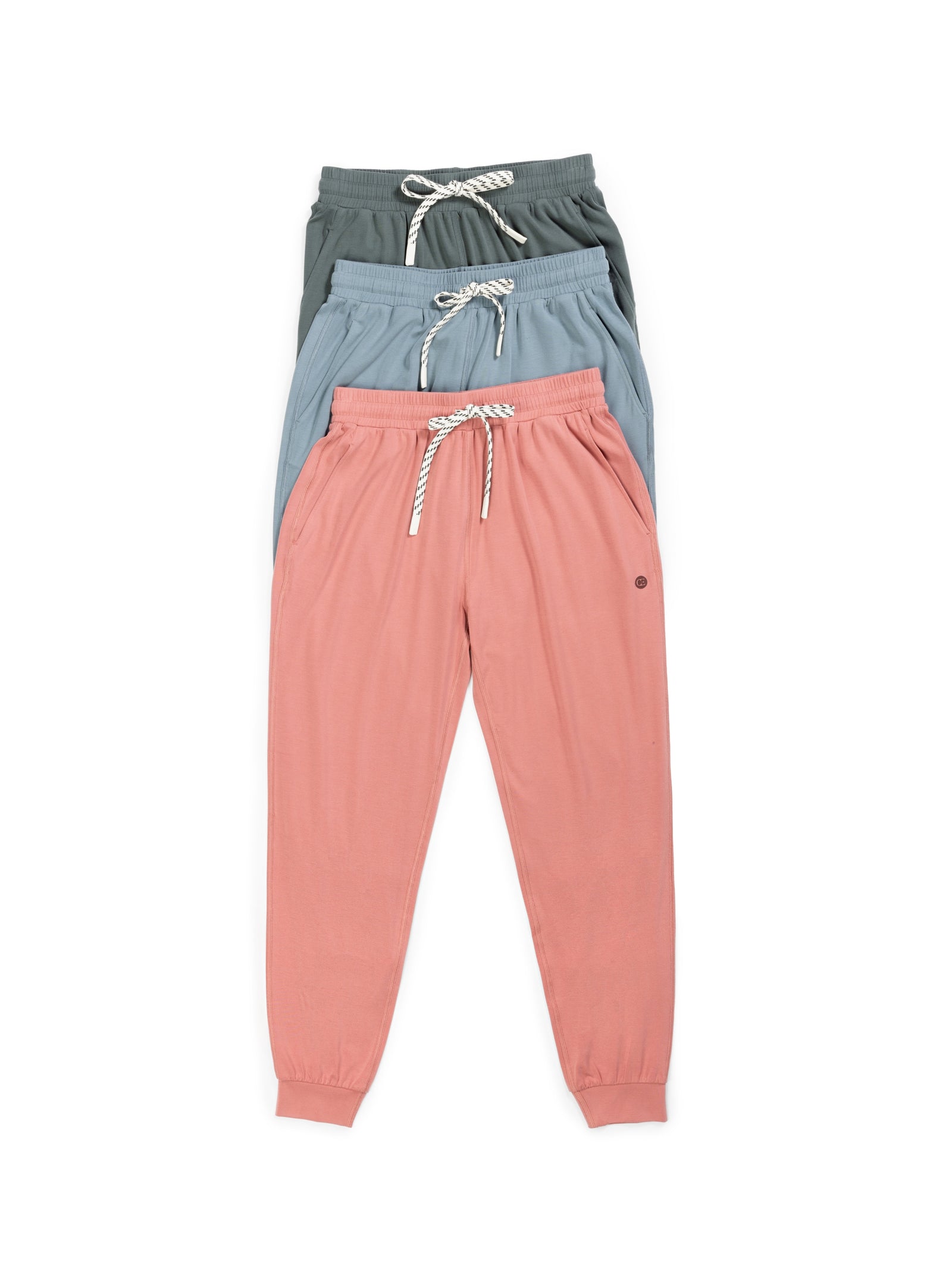 Three pairs of Women's Studio Joggers by Cozy Earth in various colors: the bottom pair in light pink, the middle pair in light blue, and the top pair in dark green. Each jogger features a white drawstring and an elastic waistband with ankle cuffs. The pants are neatly stacked on top of each other. 