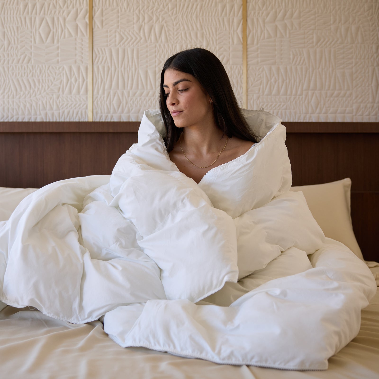 A woman, enveloped in a Cozy Earth Down Alternative Comforter, sits on a bed adorned with beige pillows. She sports long dark hair and wears a necklace. The wall behind features a textured geometric pattern, creating an overall atmosphere of coziness and serenity.