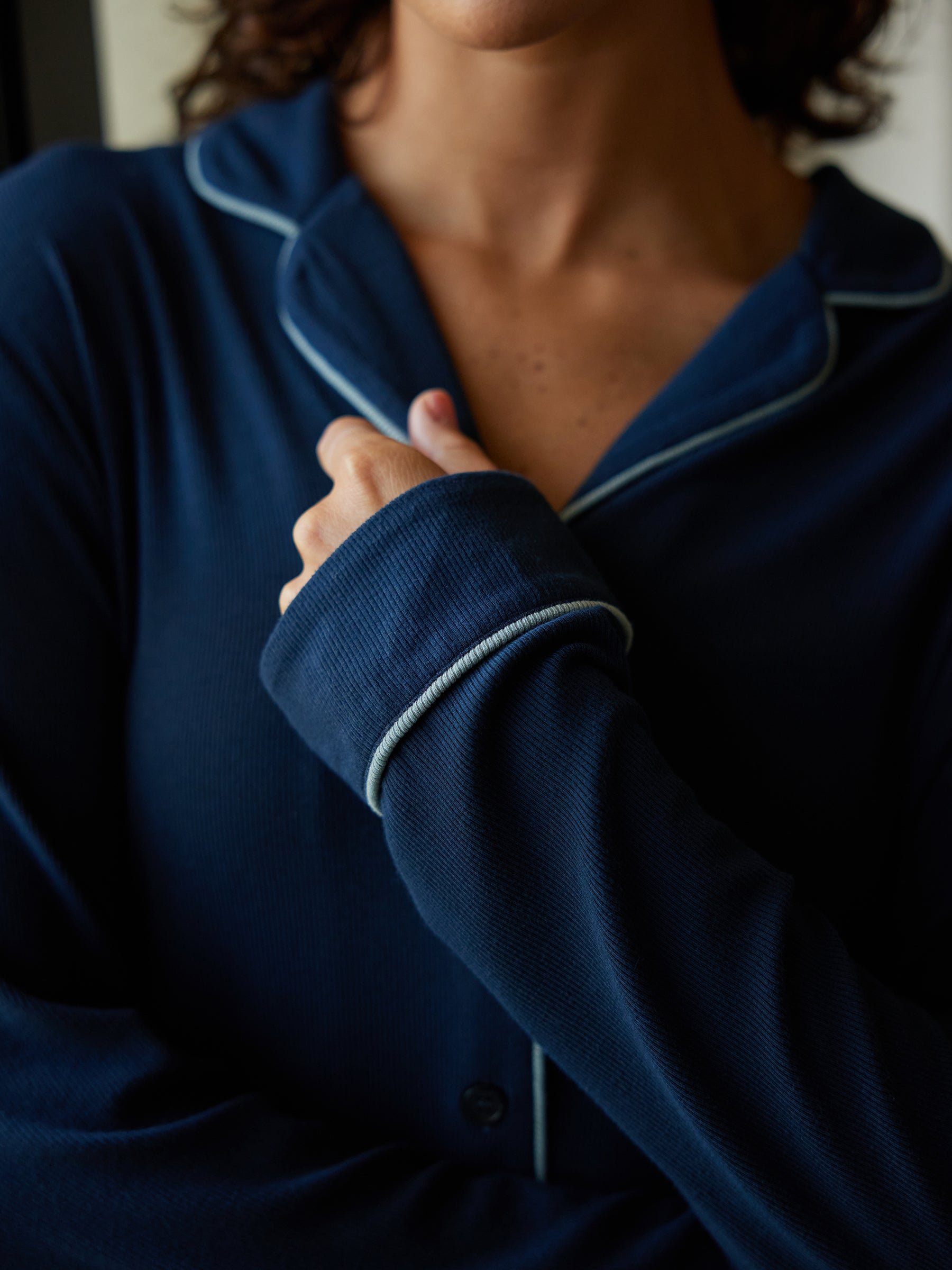 A close-up shot features an individual wearing the Women's Bamboo Rib Knit Classic Long Sleeve Pajama Top from Cozy Earth. The dark blue pajama top, accented with light blue piping detail, is being casually adjusted at the collar by the person’s hand, exuding a relaxed and comfortable vibe. The background is blurred, keeping the focus on the stylish pajamas. 