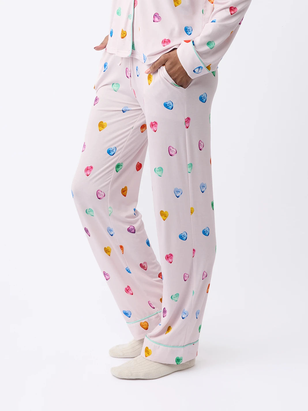 A person in Cozy Earth Women's Stretch-Knit Bamboo Pajama Pant, cream with colorful hearts, stands relaxed with hands in pockets. Light-colored socks complement the ensemble against a plain white background. |Color:Lilac Sleepy Hearts