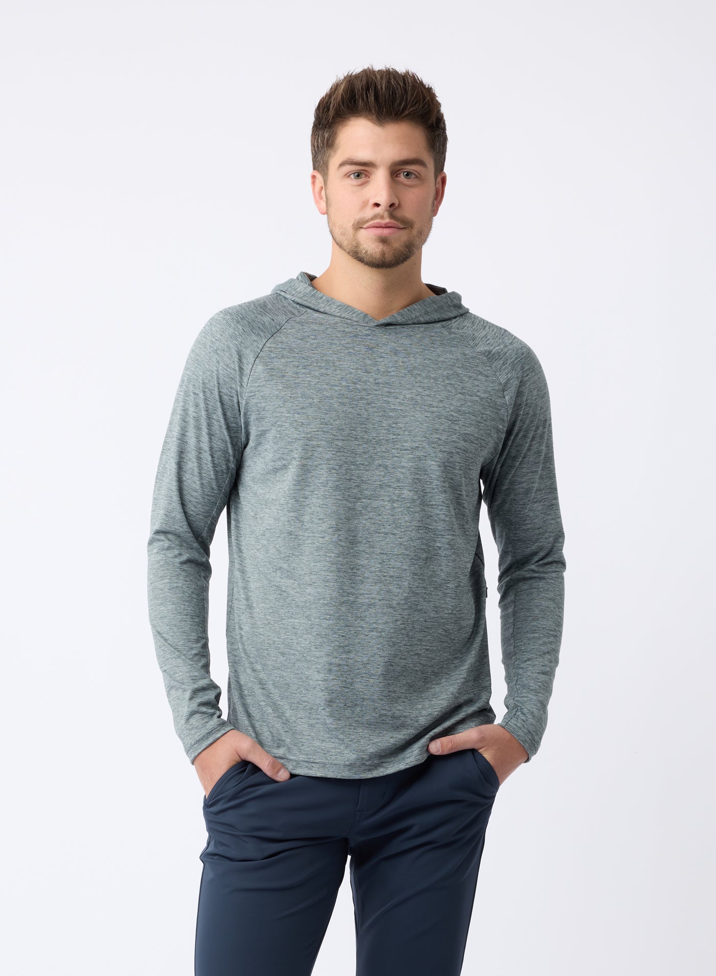 A person with short hair poses confidently against a white background, wearing Cozy Earth's Men's Lightweight Performance Hoodie in slate-gray and dark pants, with hands in pockets. |Color:Heather Dawn