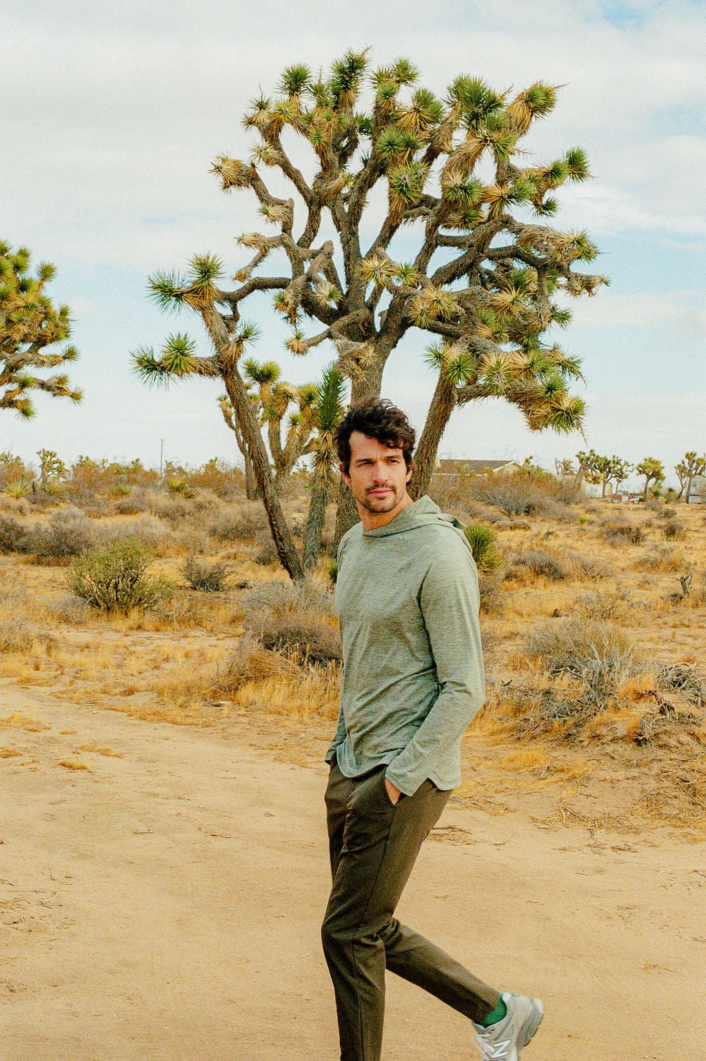 A man in a gray Men's Lightweight Performance Hoodie by Cozy Earth and dark pants strolls on a sandy desert path surrounded by Joshua trees, under a partly cloudy sky, with one hand in his pocket, looking contemplative. 