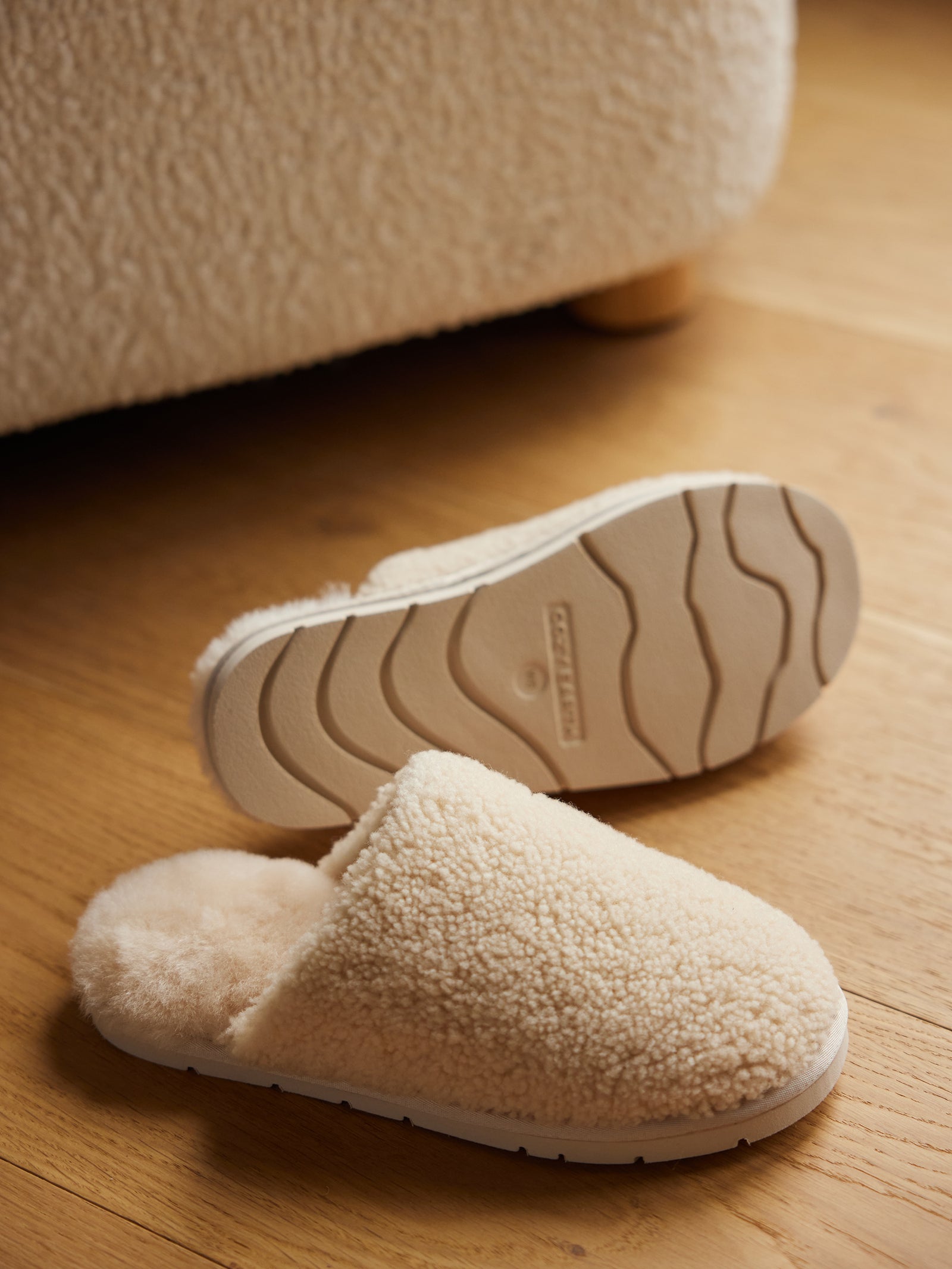 A cozy pair of Puffy Sheep Slippers from Cozy Earth, in a cream color and fluffy texture, are placed on a wooden floor. One slipper is positioned top-down to showcase its textured sole, while the other is right side up, revealing its soft, plush interior. A piece of furniture with a similar texture can be partially seen in the background. 