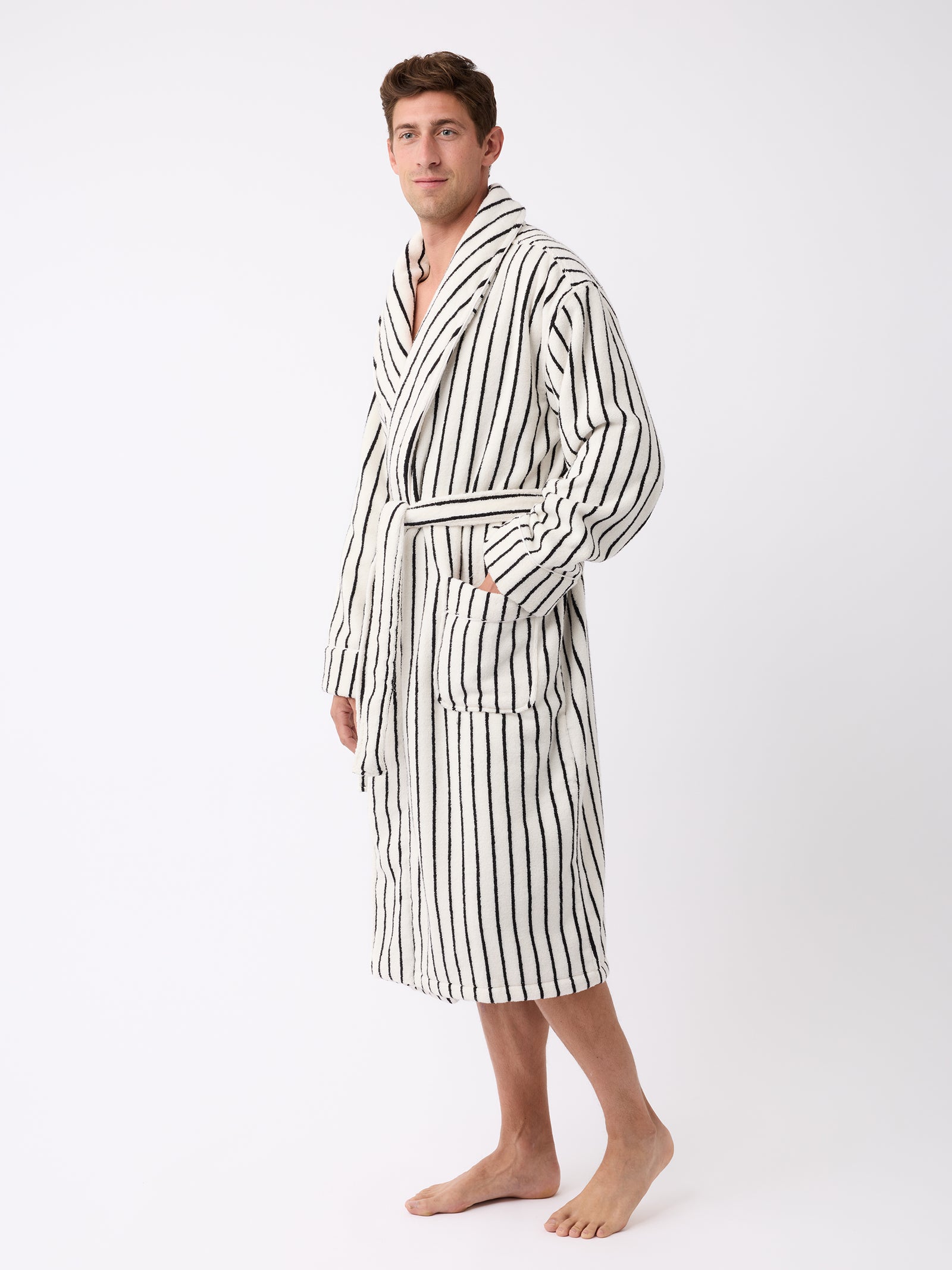 A person stands barefoot wearing the Luxe Bath Robe by Cozy Earth. The robe is white with black vertical stripes, featuring a belt and pockets. The background is plain and white. 