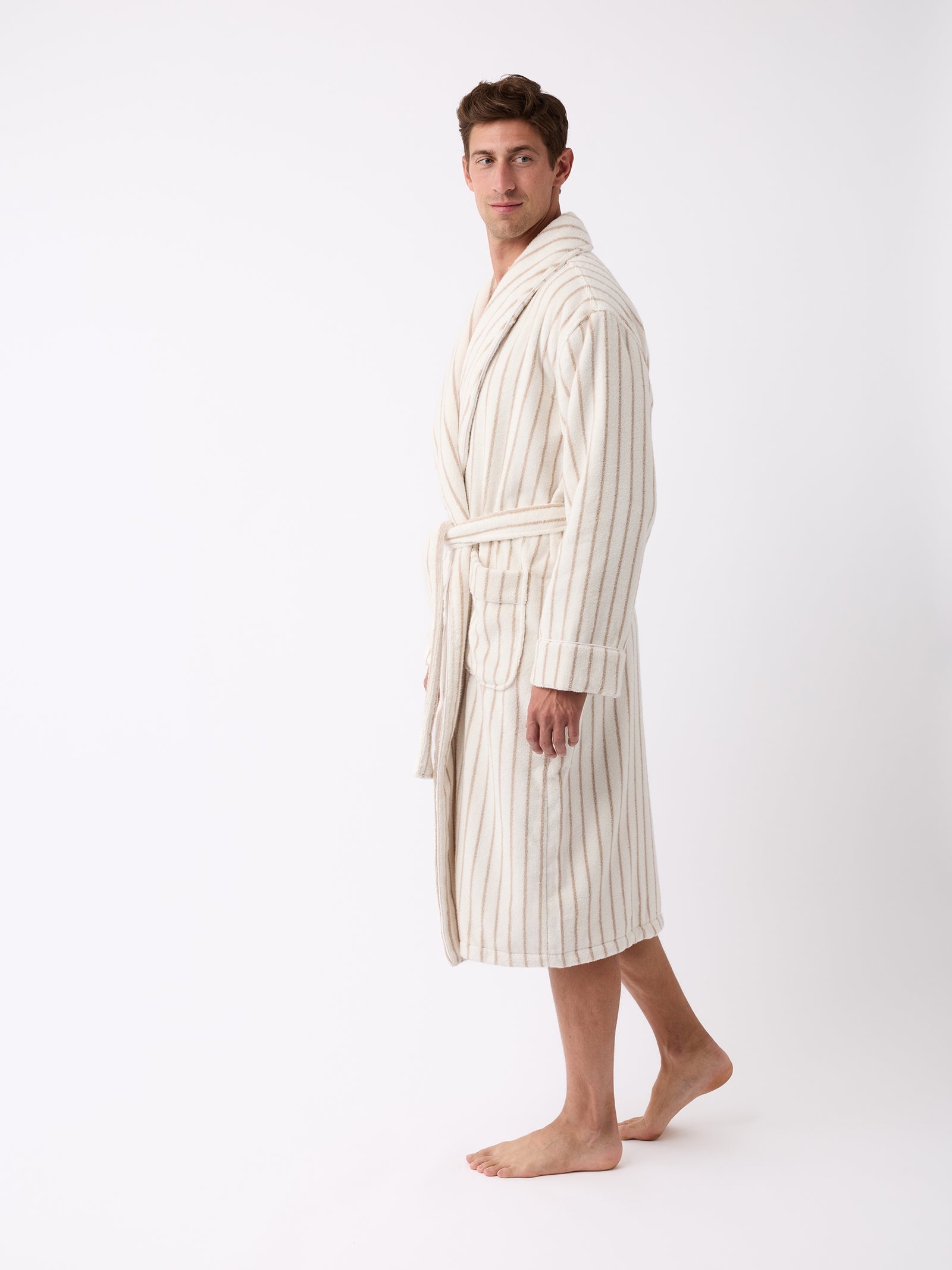 A person wearing the Luxe Bath Robe from Cozy Earth stands barefoot against a plain white background. The robe features light-colored vertical stripes, and the person gazes towards the camera with one hand in a pocket. 