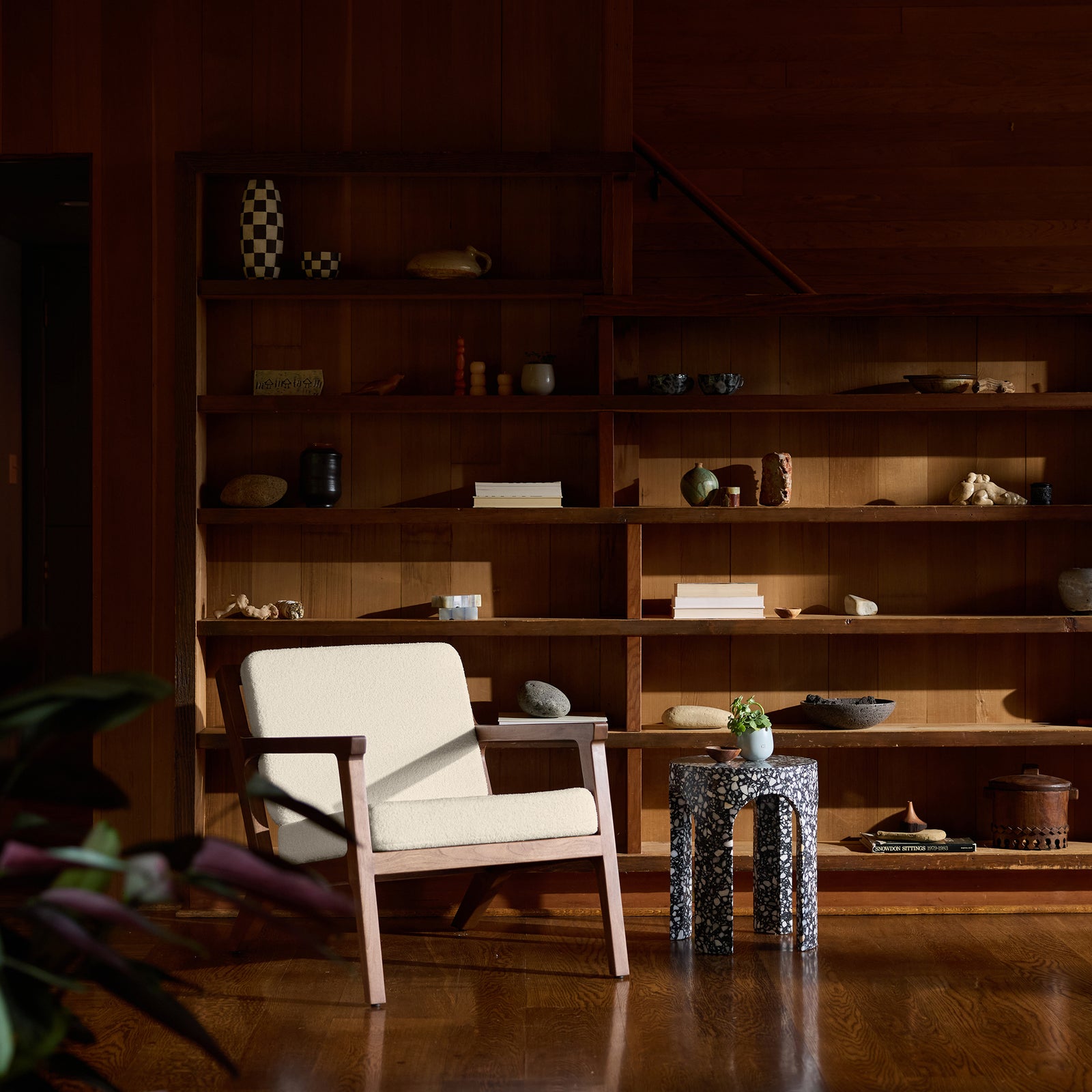 A cozy room featuring a Coronado Lounge Chair by Cozy Earth with a cream cushion, alongside a small sculptural side table and a wooden bookshelf adorned with various ceramics and decorative items. Sunlight filters in, casting warm tones on the wooden floor. 