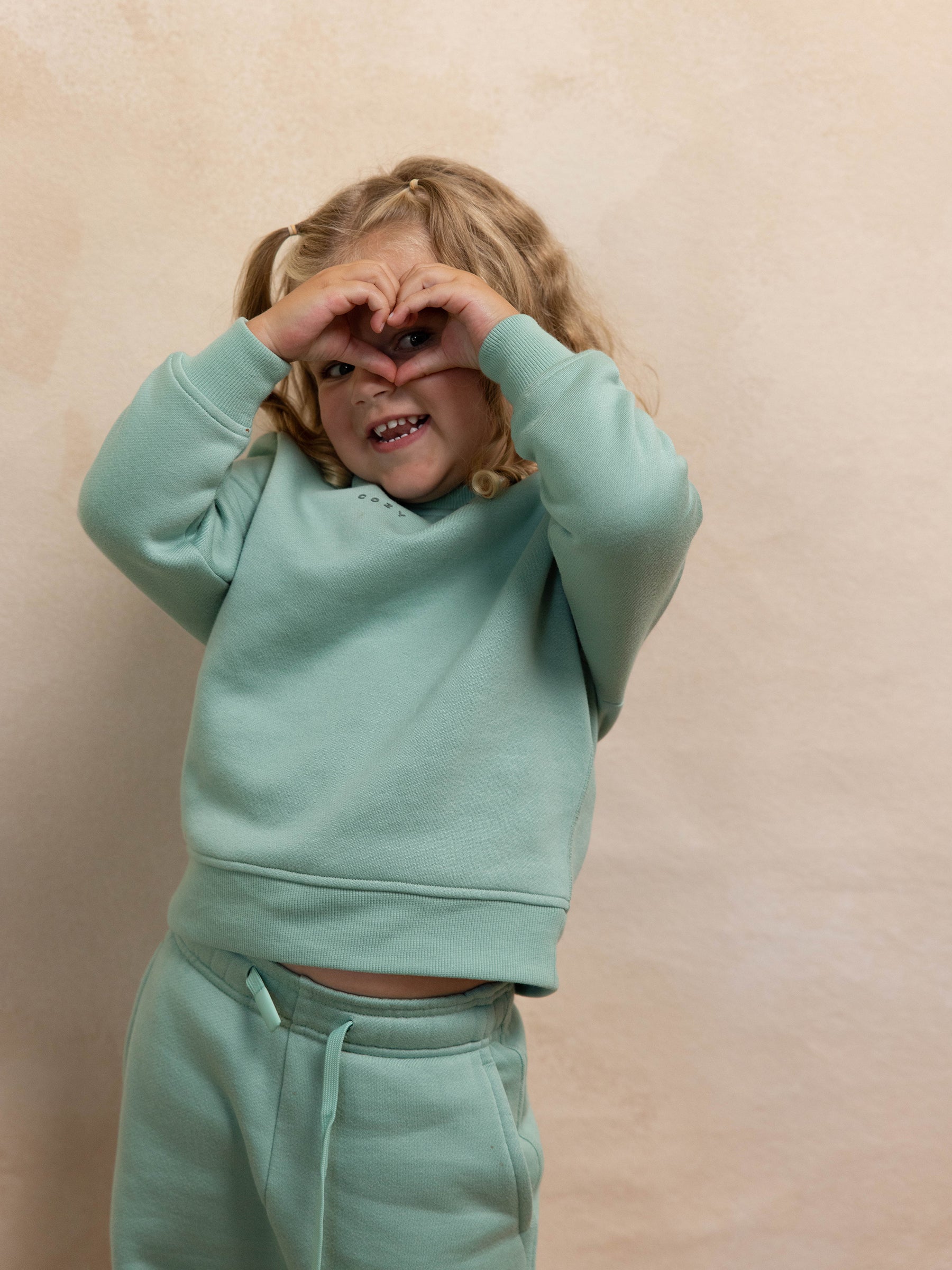 A young child with curly blonde hair wearing Cozy Earth's Kid's CityScape Crewneck in mint green forms a heart shape with their hands over their face. They are smiling and standing against a beige background. 