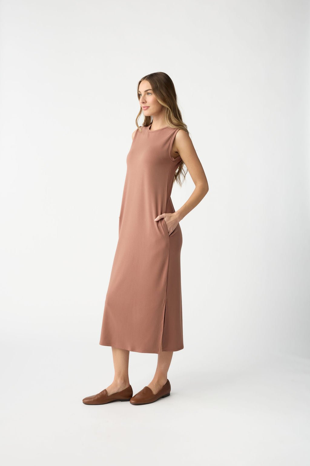 A woman stands sideways on a plain white background, wearing a Cozy Earth Women's Brushed Bamboo Sleeveless Midi Dress with pockets. The ankle-length brown dress is paired with brown slip-on shoes. Her long, wavy hair complements the look. |Color:Saddle