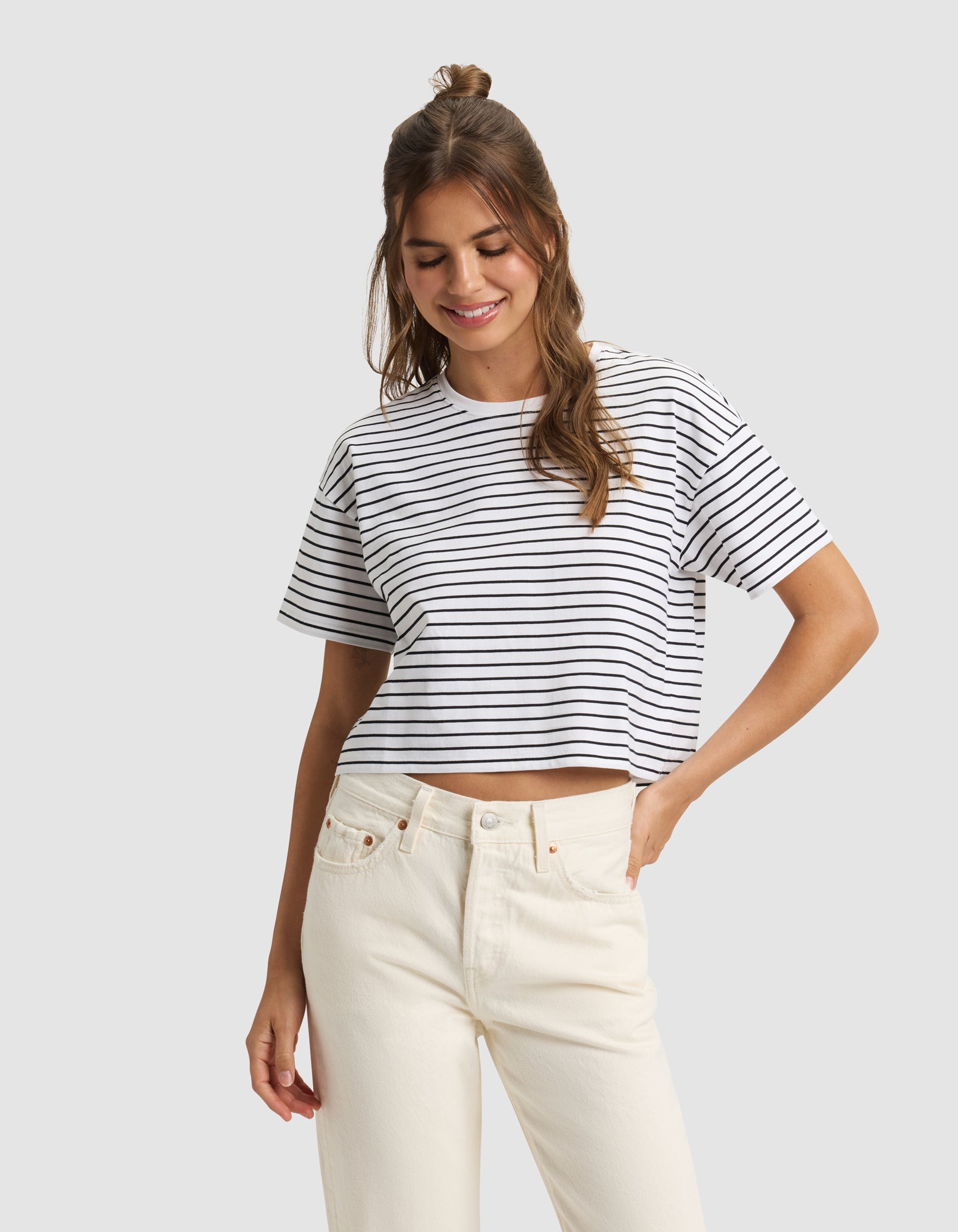 A woman with long brown hair wears the Cozy Earth Women's All Day Cropped Tee, featuring white and black stripes, and high-waisted cream jeans, standing against a light gray background with a smile and one hand in her pocket. 