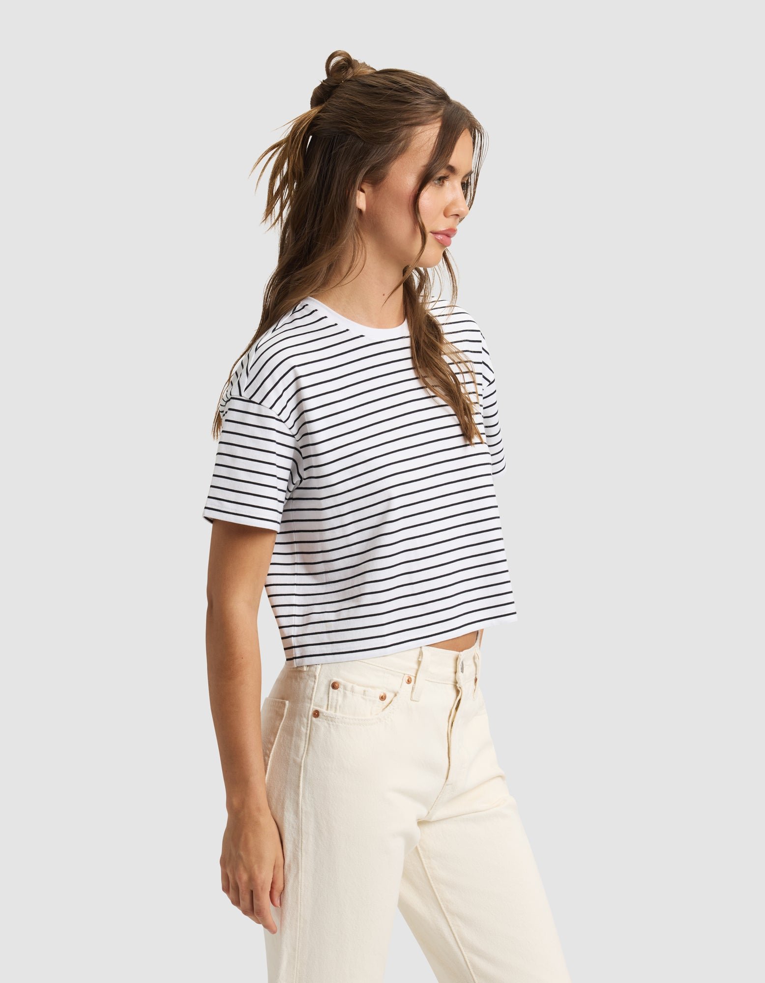A woman with long, wavy brown hair wears the Cozy Earth Women's All Day Cropped Tee in white and navy stripes with high-waisted off-white jeans, standing in profile against a plain light background. |Color:Jet Black Stripe