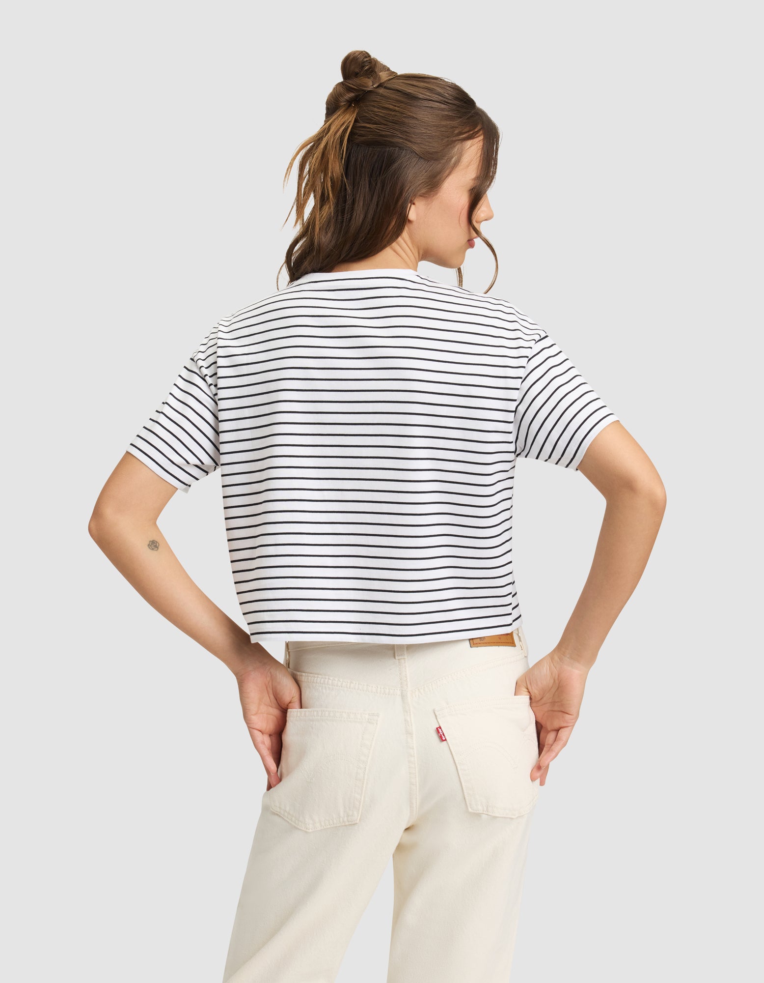 A person stands with their back to the camera, wearing Cozy Earth's Women's All Day Cropped Tee in white and black stripes and off-white jeans. Their hands are on their hips, brown hair half-up, against a plain light gray background. 