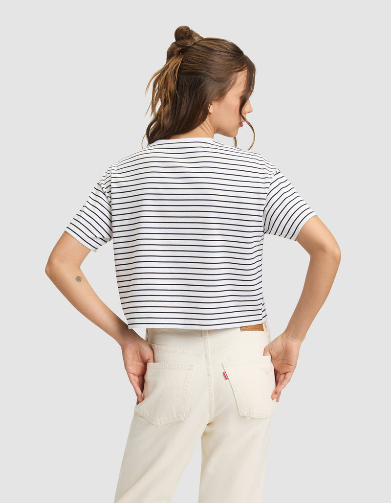 A person stands with their back to the camera, wearing Cozy Earth's Women's All Day Cropped Tee in white and black stripes and off-white jeans. Their hands are on their hips, brown hair half-up, against a plain light gray background.