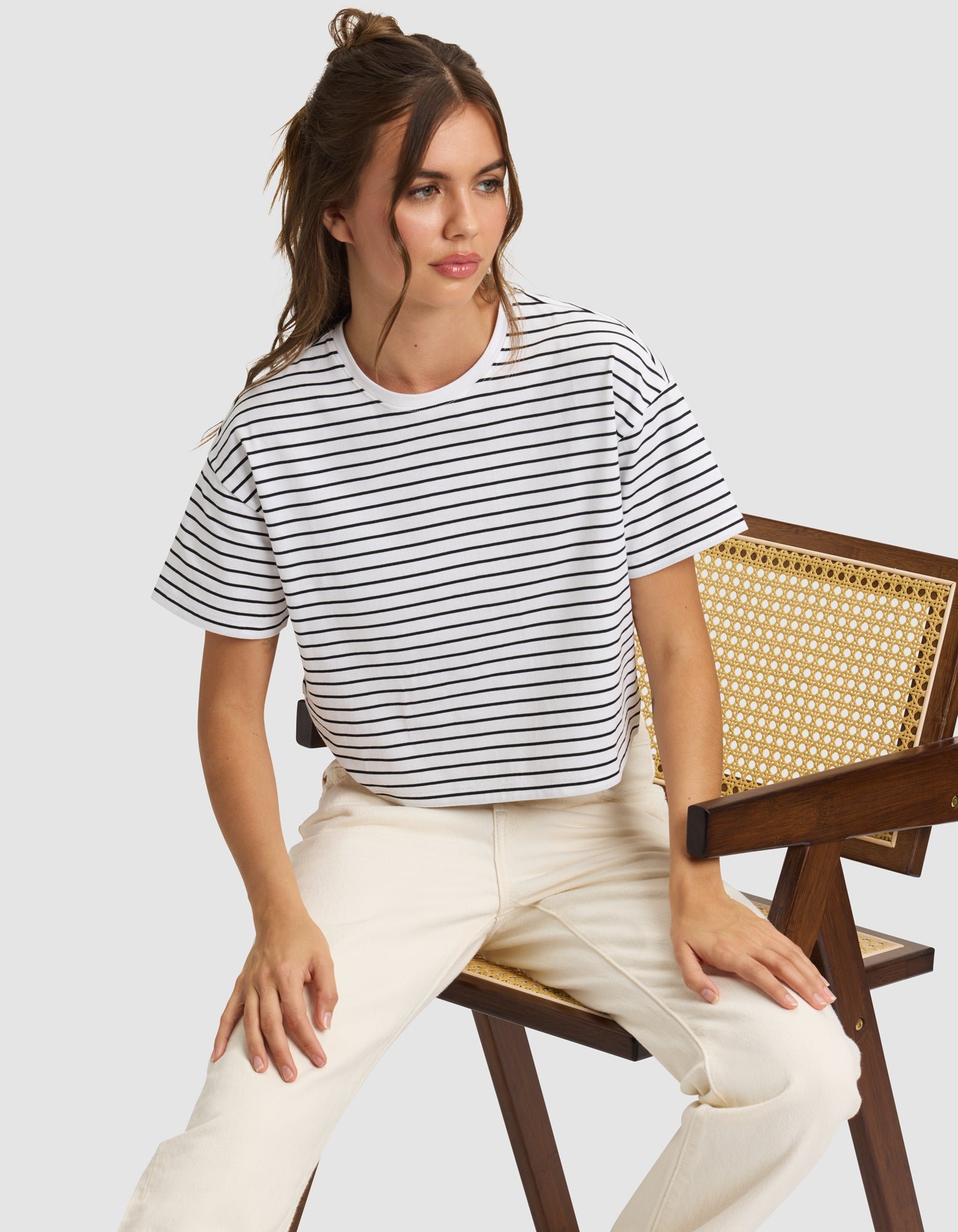 A woman with long brown hair in a half-up style sits on a wooden chair, wearing the Cozy Earth Women's All Day Cropped Tee in white with black stripes and off-white pants, looking slightly to the side. |Color:Jet Black Stripe