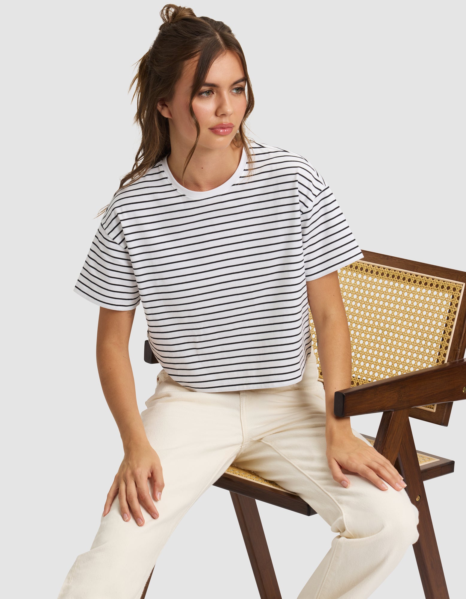 A woman with long brown hair in a half-up style sits on a wooden chair, wearing the Cozy Earth Women's All Day Cropped Tee in white with black stripes and off-white pants, looking slightly to the side. 