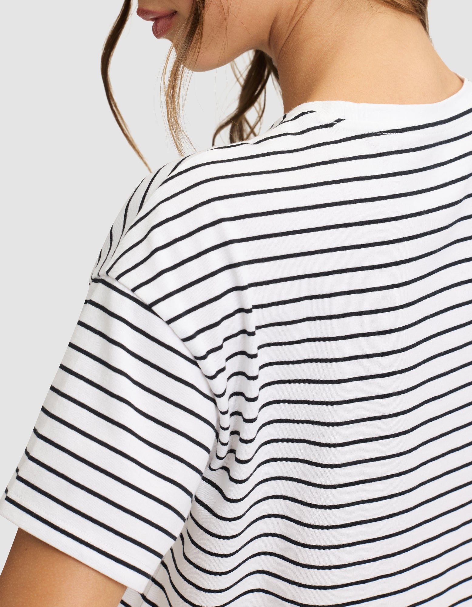 A close-up shows a person wearing the Cozy Earth Women's All Day Cropped Tee, a white top with thin black horizontal stripes, viewed from the back and side with part of their face and hair visible. 