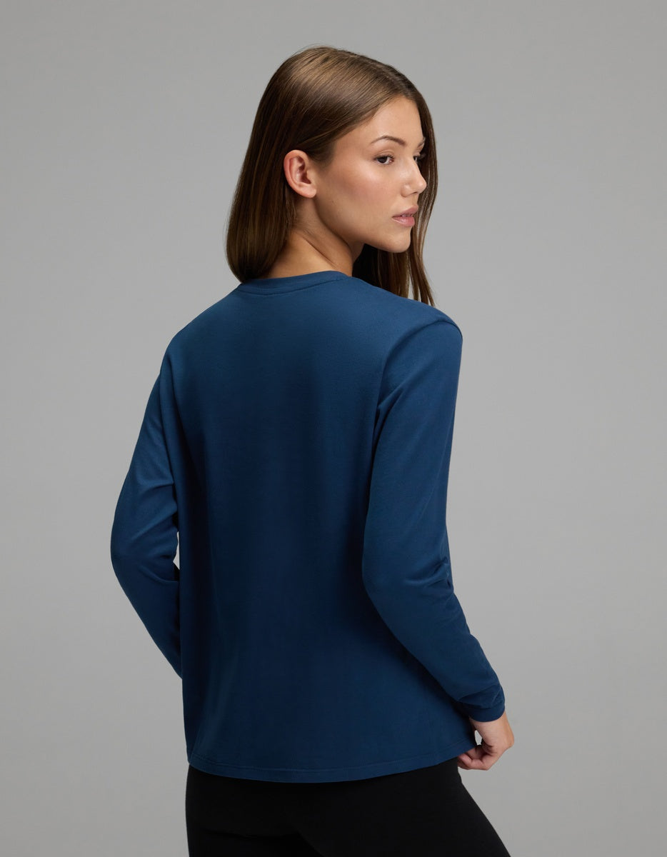 A woman with straight brown hair stands with her back to the camera and her head turned slightly over her left shoulder against a plain gray background, wearing the Cozy Earth Women’s All Day Long Sleeve Tee in dark blue paired with black pants.