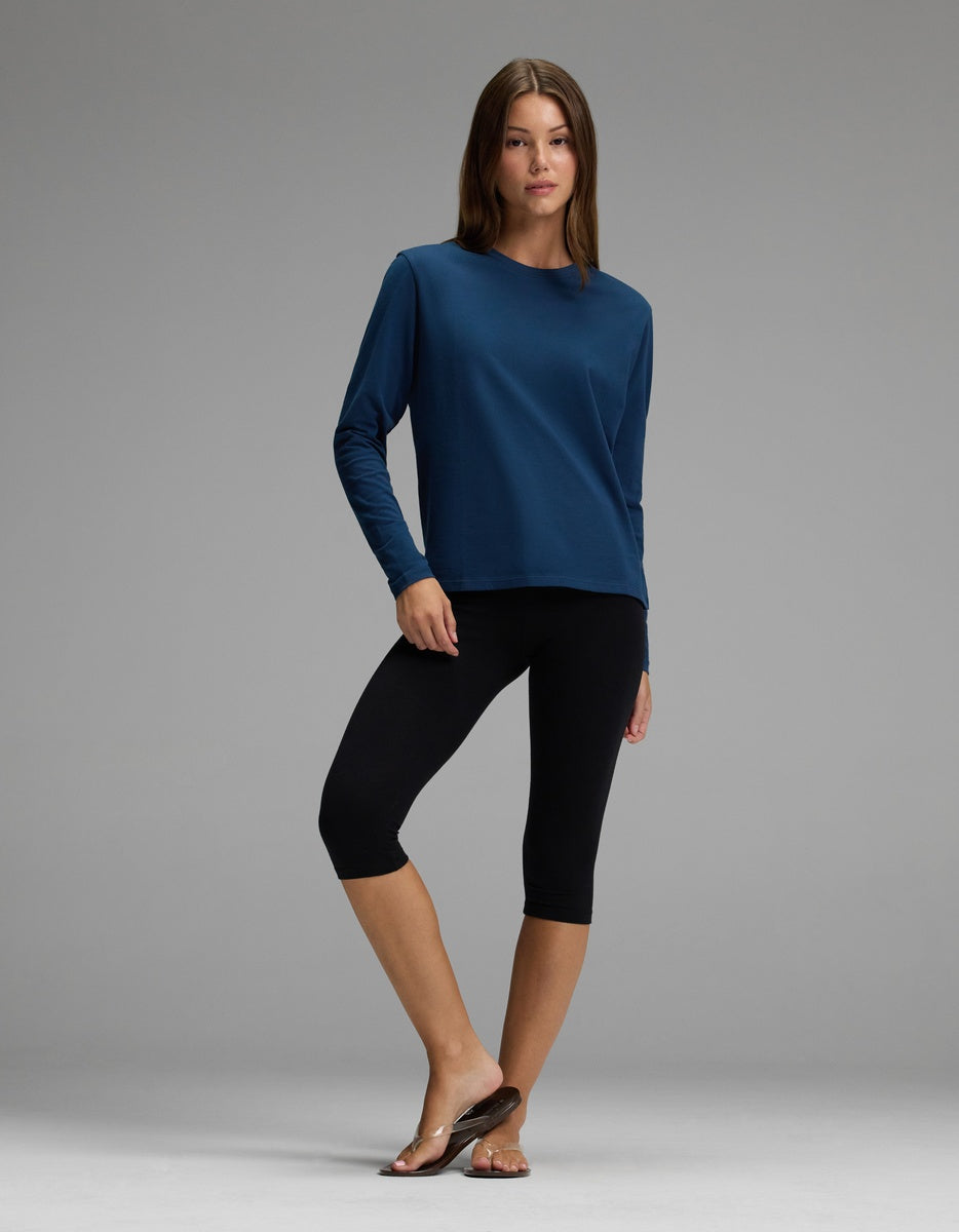A woman stands against a gray background wearing the Cozy Earth Women’s All Day Long Sleeve Tee in blue, paired with black capri leggings and brown sandals. One leg is slightly raised, with her hand resting on her thigh.