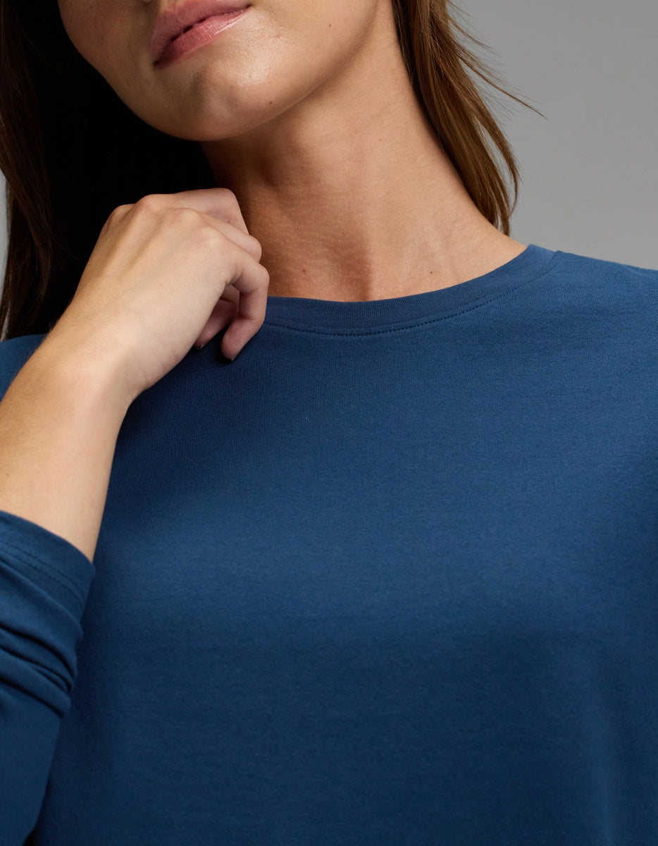 A woman wearing the Cozy Earth Women’s All Day Long Sleeve Tee in blue gently touches her neck with one hand. Her face is partially visible, set against a plain gray background.