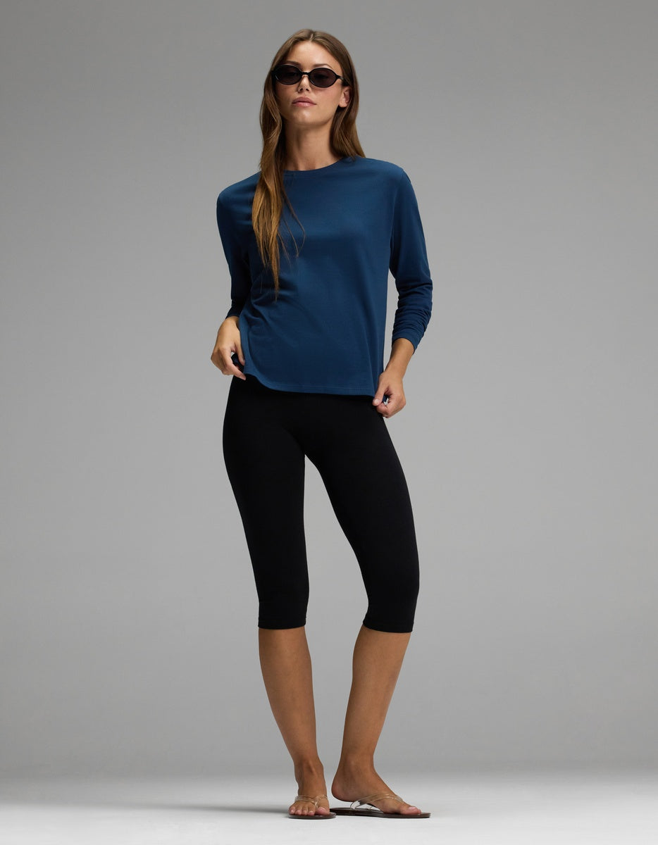 A person wearing the Cozy Earth Women’s All Day Long Sleeve Tee in blue stands barefoot against a plain gray background, paired with black knee-length leggings and sunglasses.