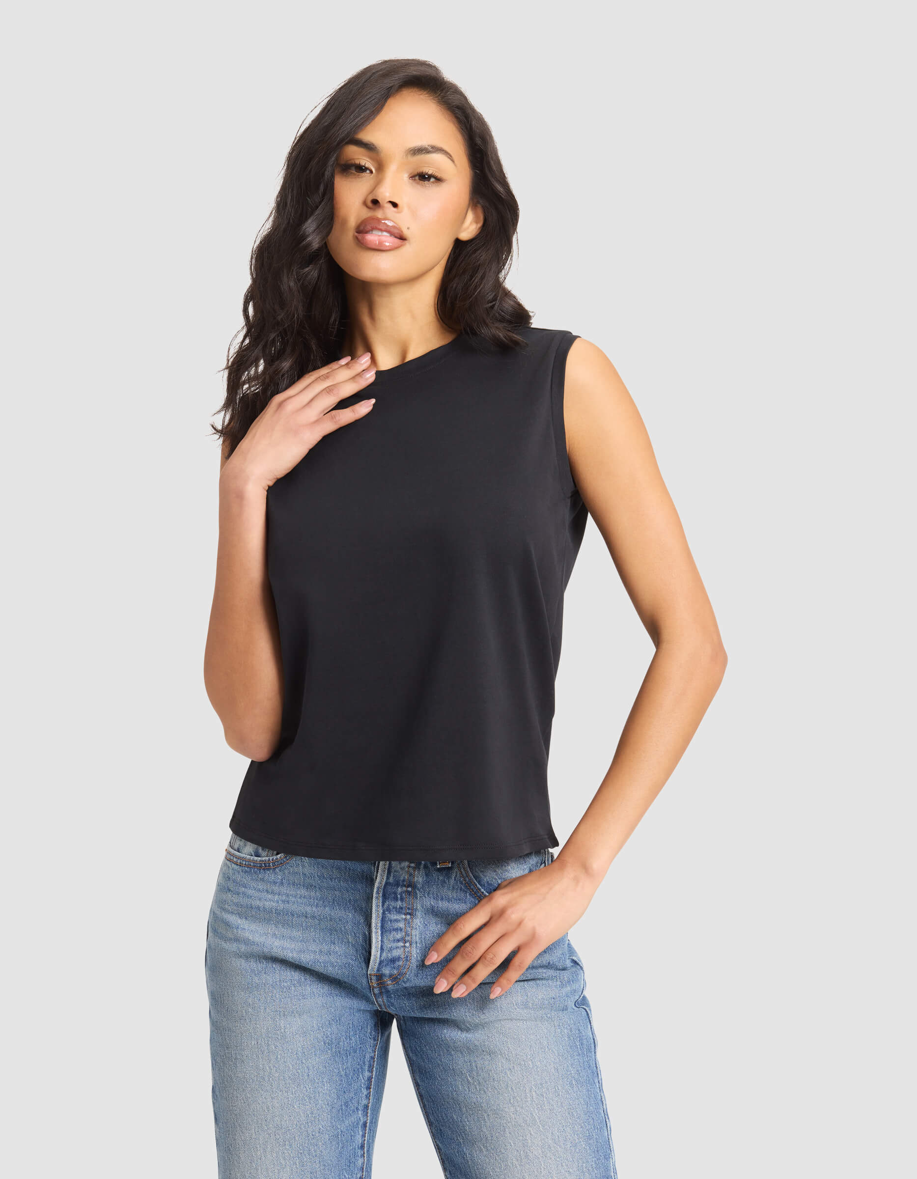 A woman models the Cozy Earth Women's All Day Sleeveless Tee in black with light blue jeans, posing against a plain light gray background.|Color:Jet Black