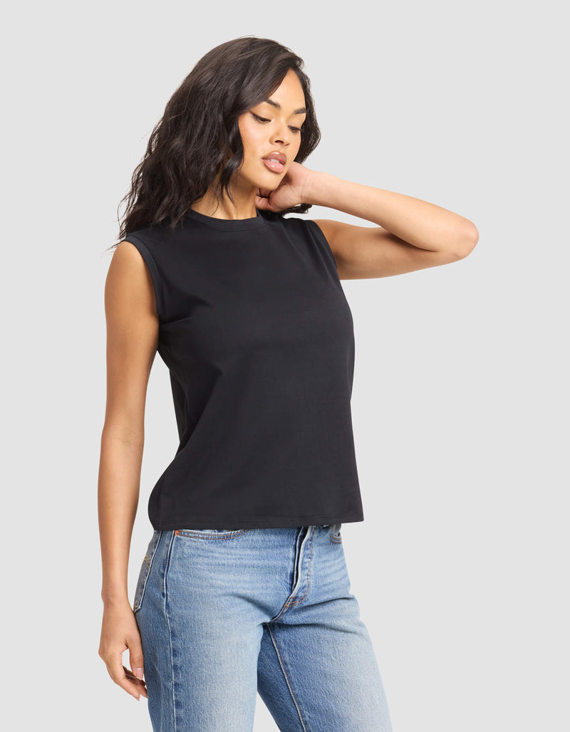 A woman with wavy dark hair models the Cozy Earth Women's All Day Sleeveless Tee in black, paired with light blue jeans. She stands against a plain light gray background, touching her hair and looking down.