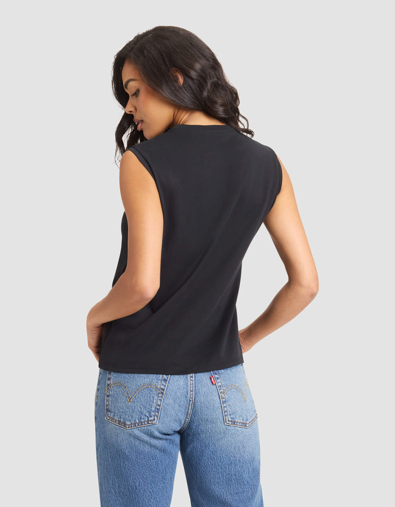 A woman with wavy dark hair models the Cozy Earth Women's All Day Sleeveless Tee in black, paired with blue jeans, standing with her back to the camera and looking over her shoulder against a plain light background.