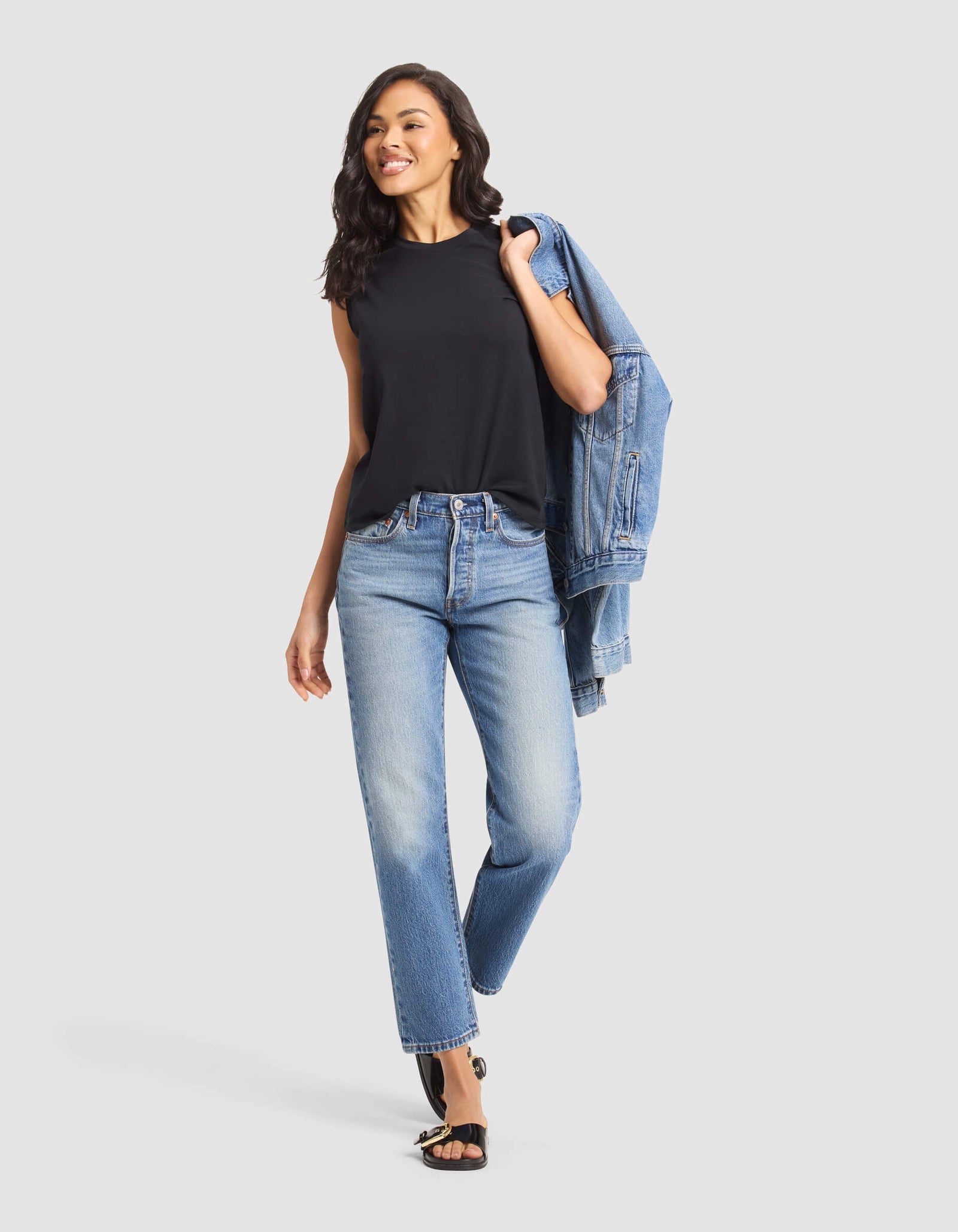 A woman smiles while walking against a plain light background, wearing Cozy Earth's Women's All Day Sleeveless Tee in black, paired with light blue jeans, black sandals, and carrying a blue denim jacket over her shoulder.
