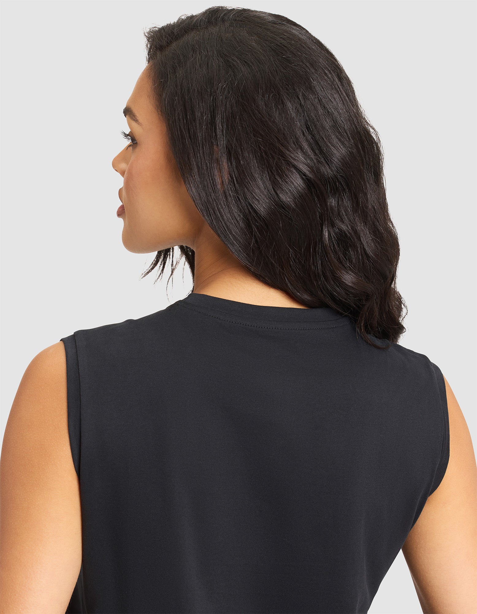 A woman with wavy dark hair is seen from behind and slightly to the side, wearing the Cozy Earth Women's All Day Sleeveless Tee in black against a plain light gray background. 