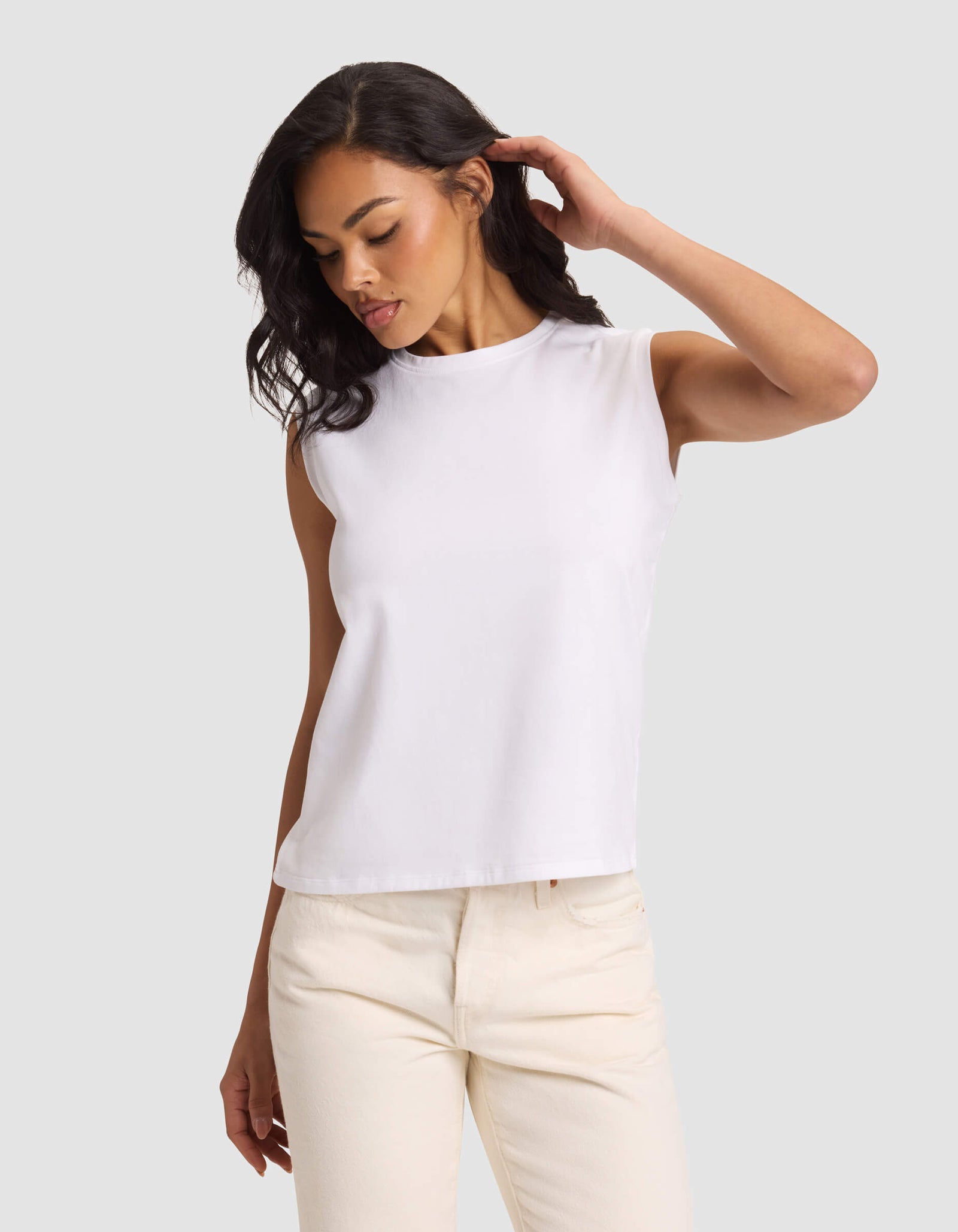 A woman models the Cozy Earth Women's All Day Sleeveless Tee in white, paired with light beige pants, standing against a plain light gray background and looking down with one hand touching her hair.