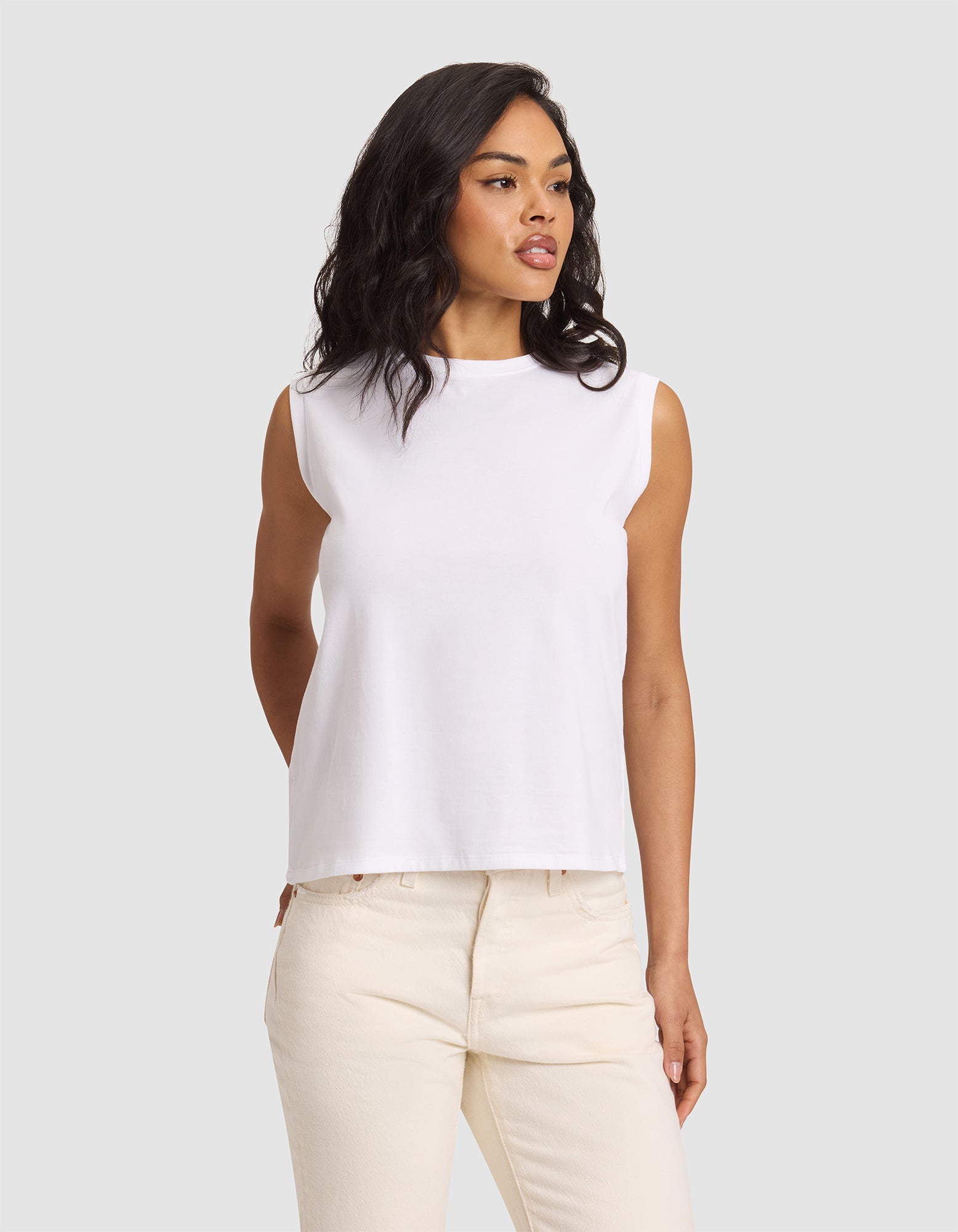 A woman with wavy dark hair models the Cozy Earth Women's All Day Sleeveless Tee in white, standing against a plain light gray background and gazing slightly to the side.