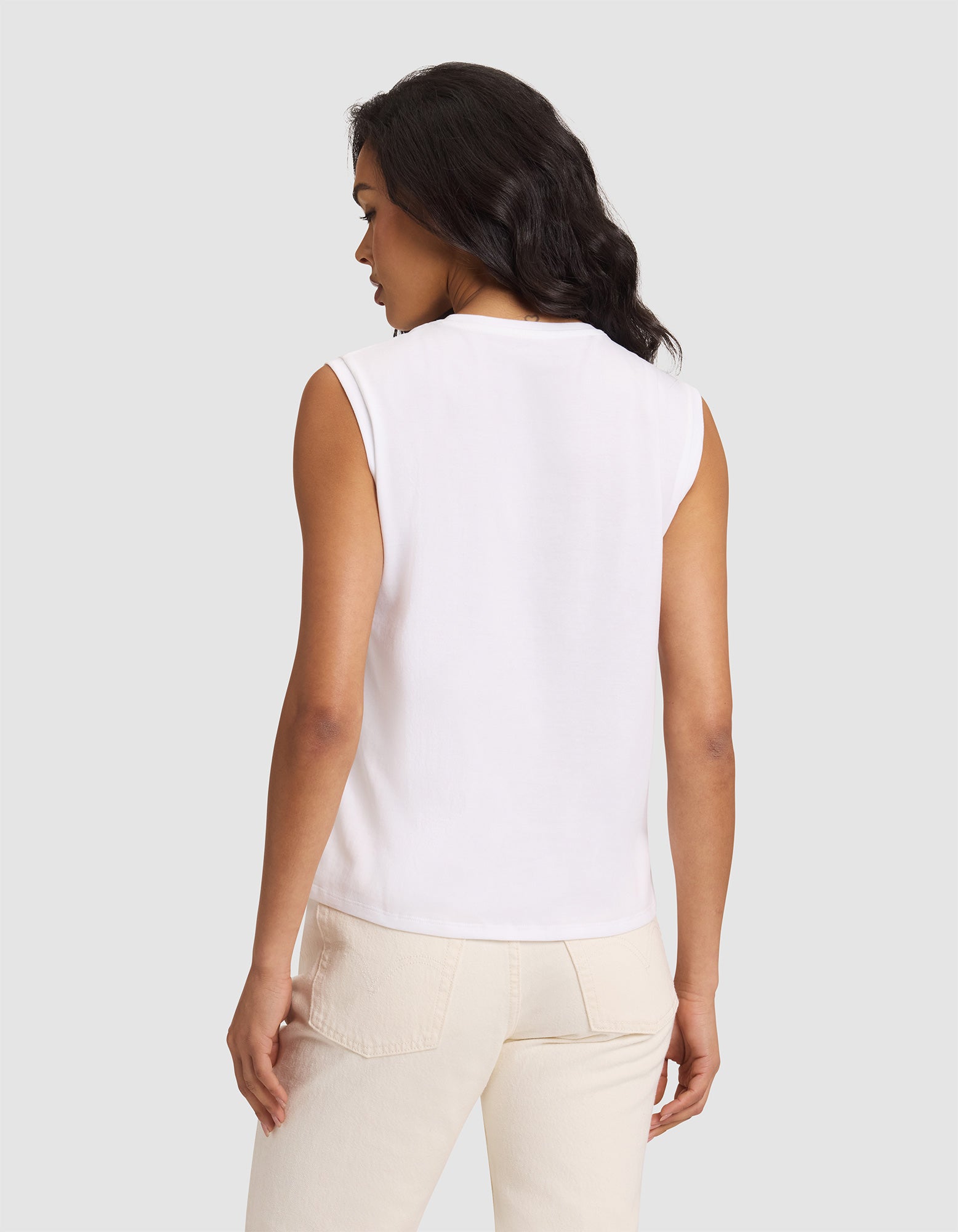 A woman with long dark hair, seen from the back, wears a Cozy Earth Women's All Day Sleeveless Tee in white and light beige pants, standing against a plain light gray background. 