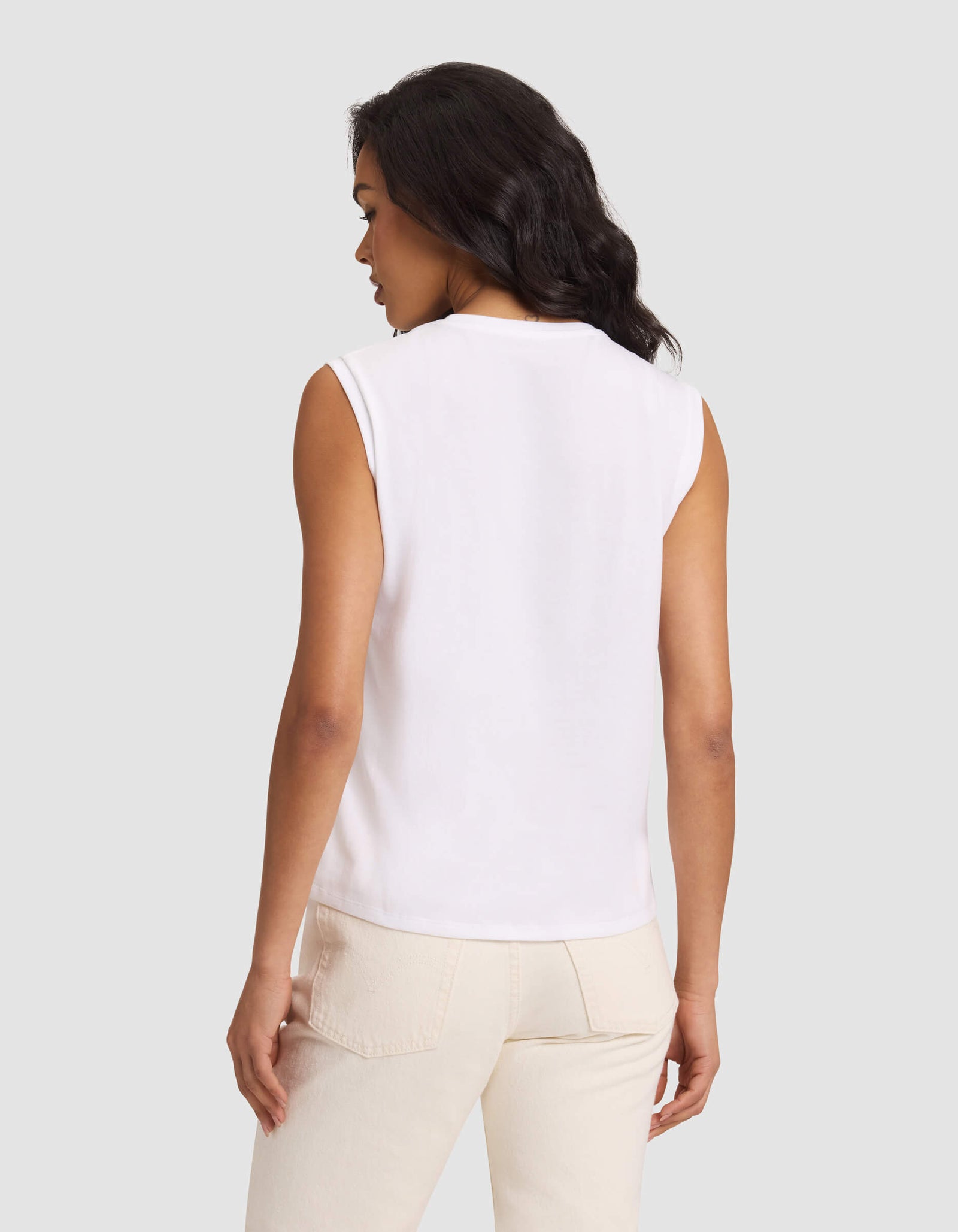 A woman with long dark hair, seen from behind, wears the Cozy Earth Women's All Day Sleeveless Tee in white and light beige pants, standing against a plain light gray background.
