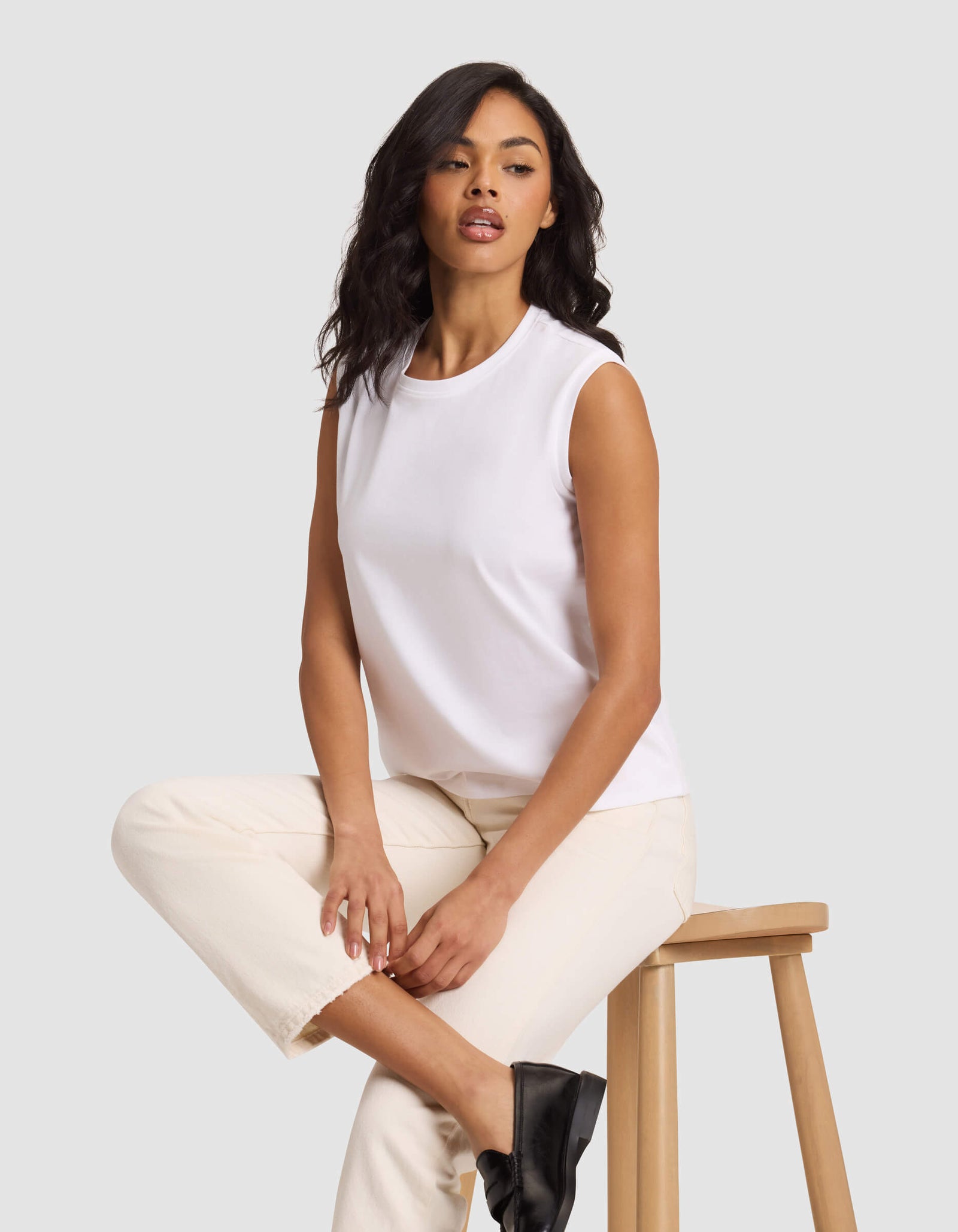 A woman with long, wavy dark hair sits on a wooden stool against a light gray background, wearing Cozy Earth's Women's All Day Sleeveless Tee in white, paired with cream pants and black shoes.