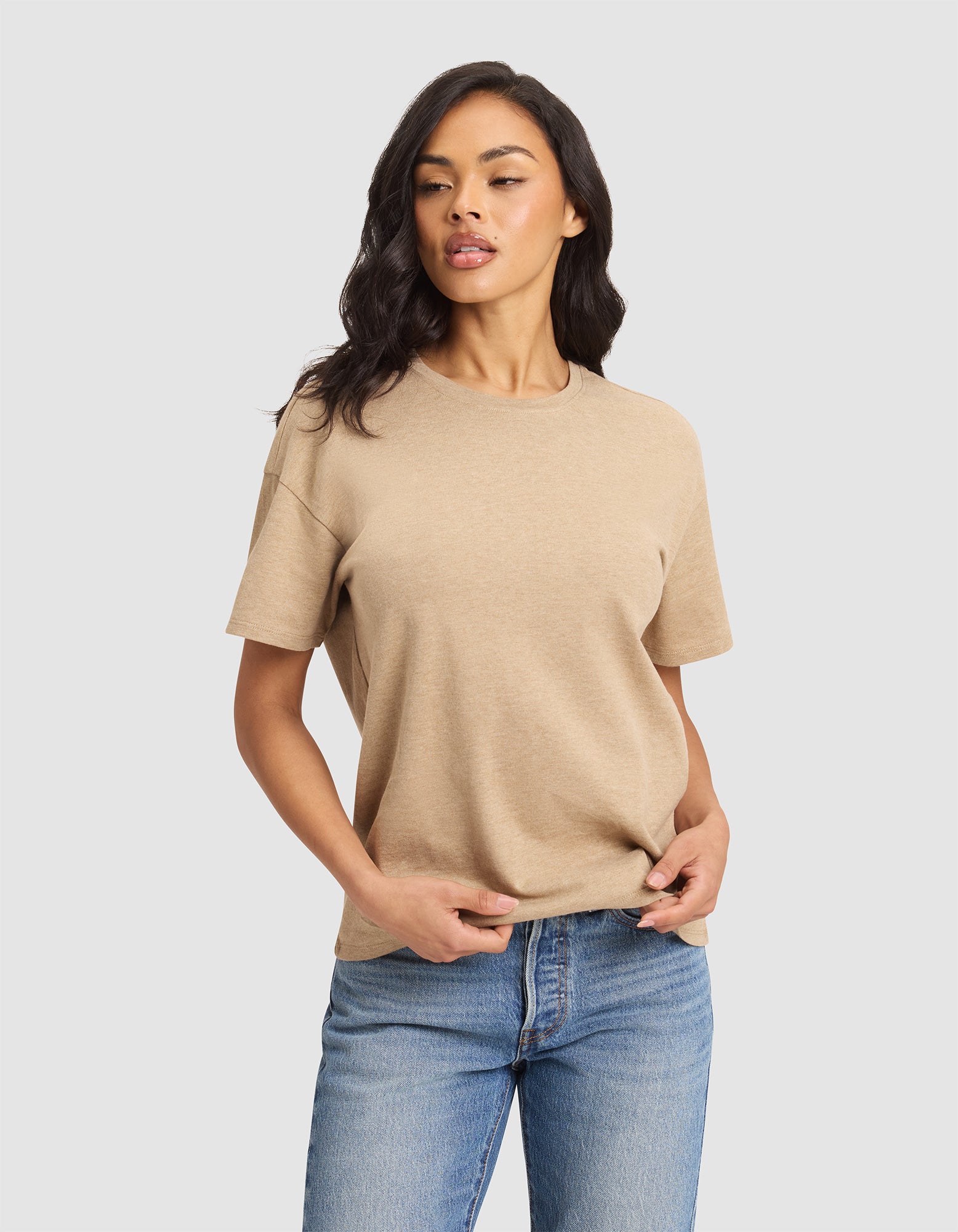 A woman with long dark hair models the Cozy Earth Women's All Day Tee in beige, paired with blue jeans, against a plain light gray background. She looks slightly to the side and holds the hem of her shirt with one hand. |Color:Beech Heather