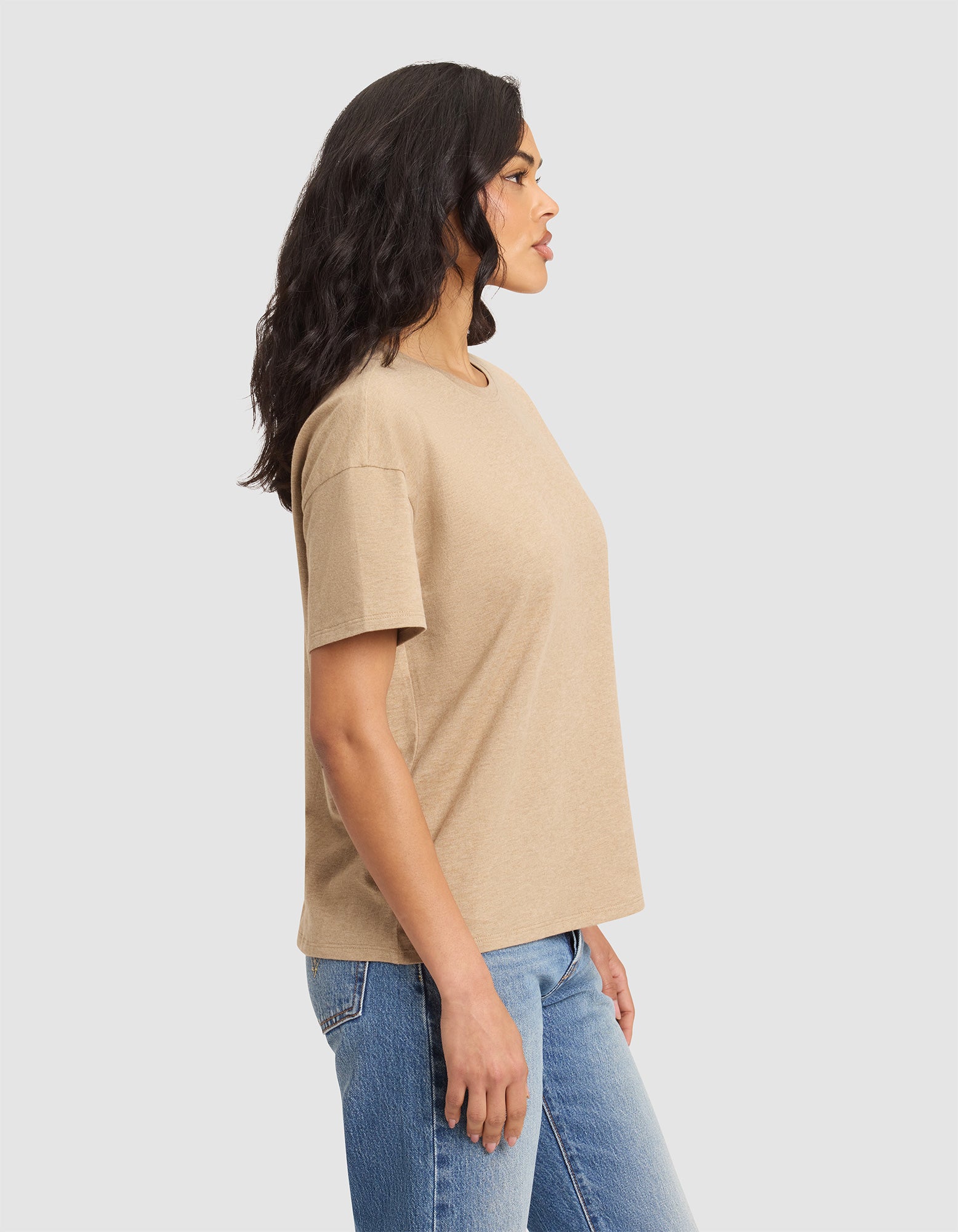 A woman with wavy dark hair stands in profile, wearing Cozy Earth's Women's All Day Tee in beige and blue jeans, set against a plain light gray background. |Color:Beech Heather